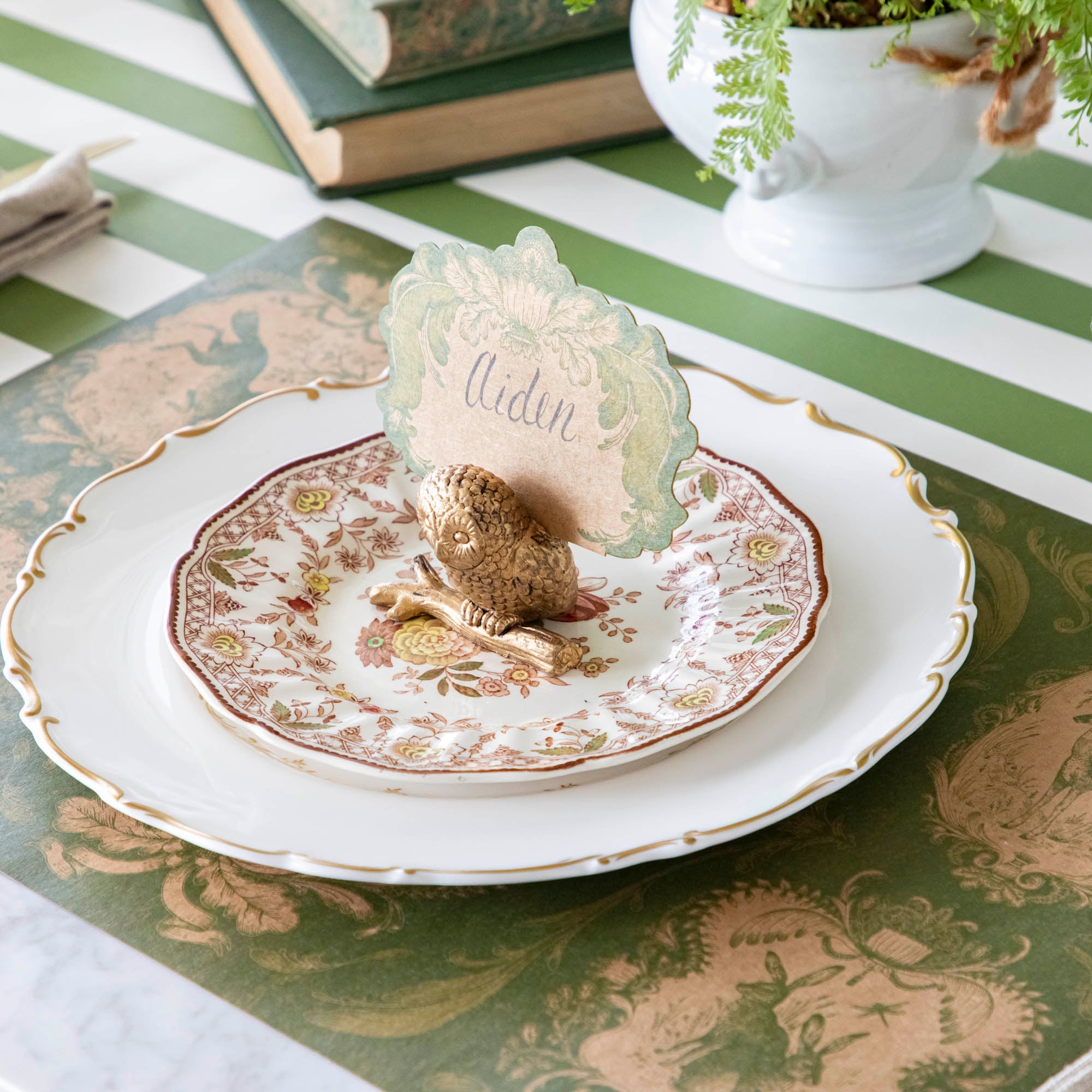 The Owl Name Card Holder holding the Moss Fable Toile Place Card with "Aiden" written on it in cursive, resting on two dinner plates with the Moss Fable Toile Placemat and Moss Green Classic Stripe Runner underneath.