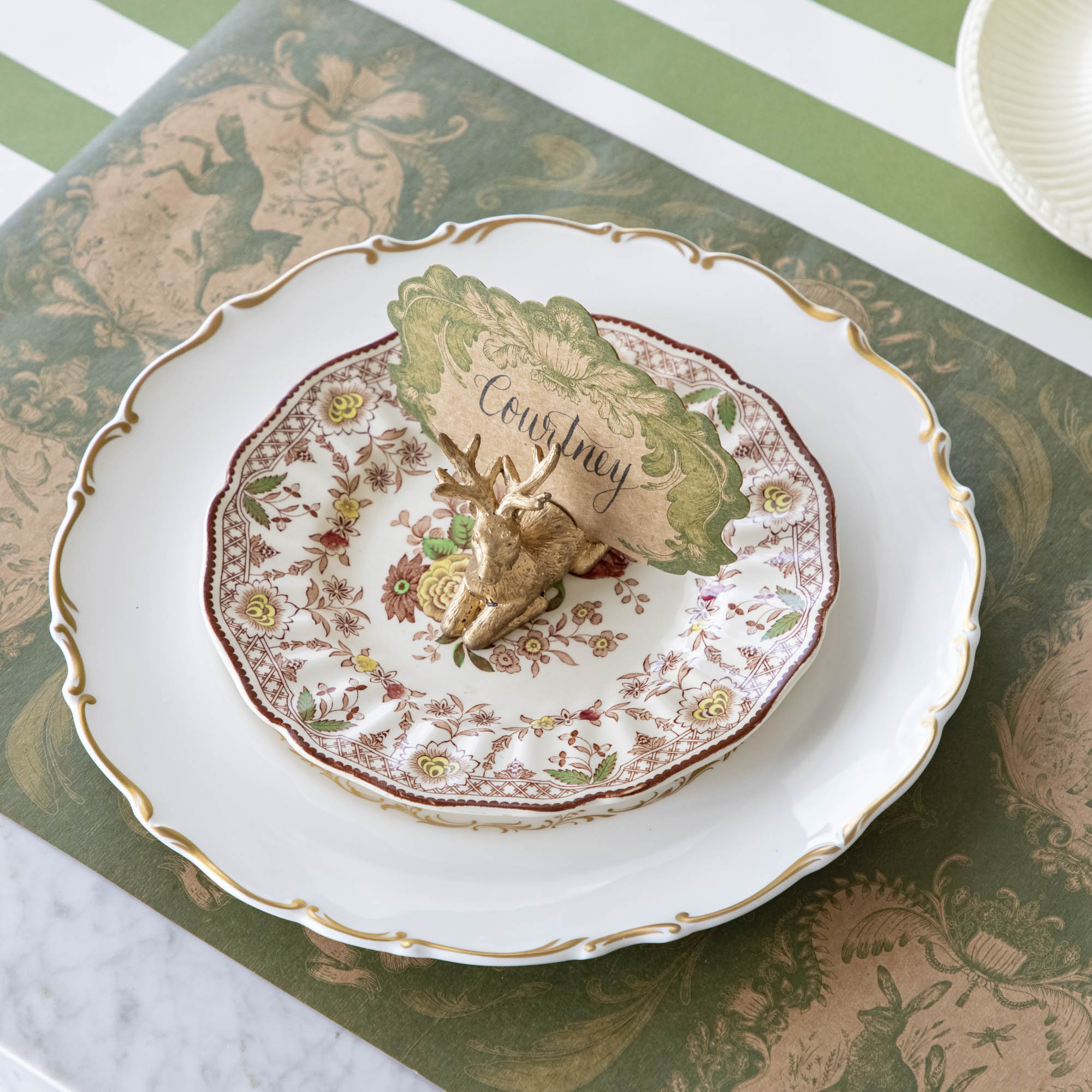 The Deer Name Card holder holding the Moss Fable Toile Place Card with "Courtney" written on it in cursive, resting on dinner plates with the Moss Fable Toile Placemat underneath.