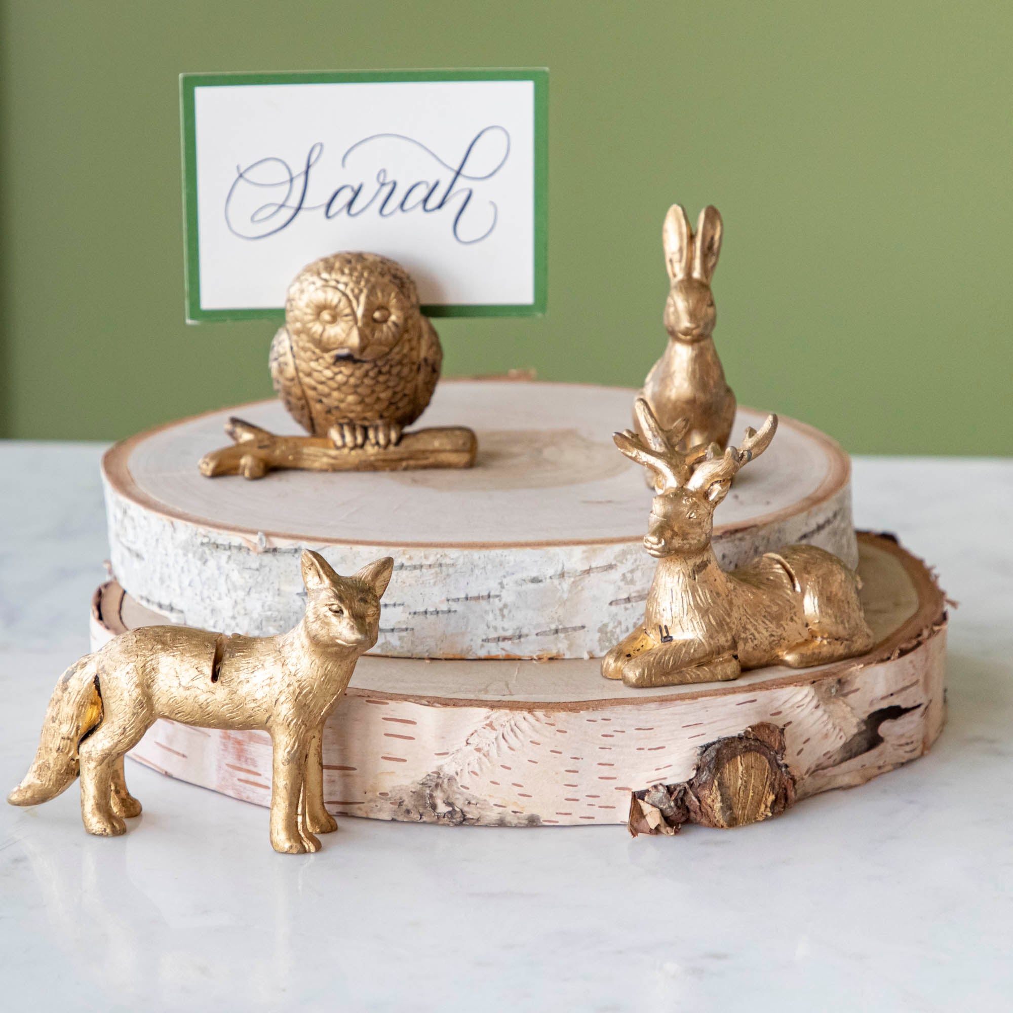 An Owl, Rabbit, Fox and Deer place card holder stacked on cylinders of wood with the Dark Green Frame Place Card with "Sarah" written on it in cursive, being held up by a slot in the owl&
