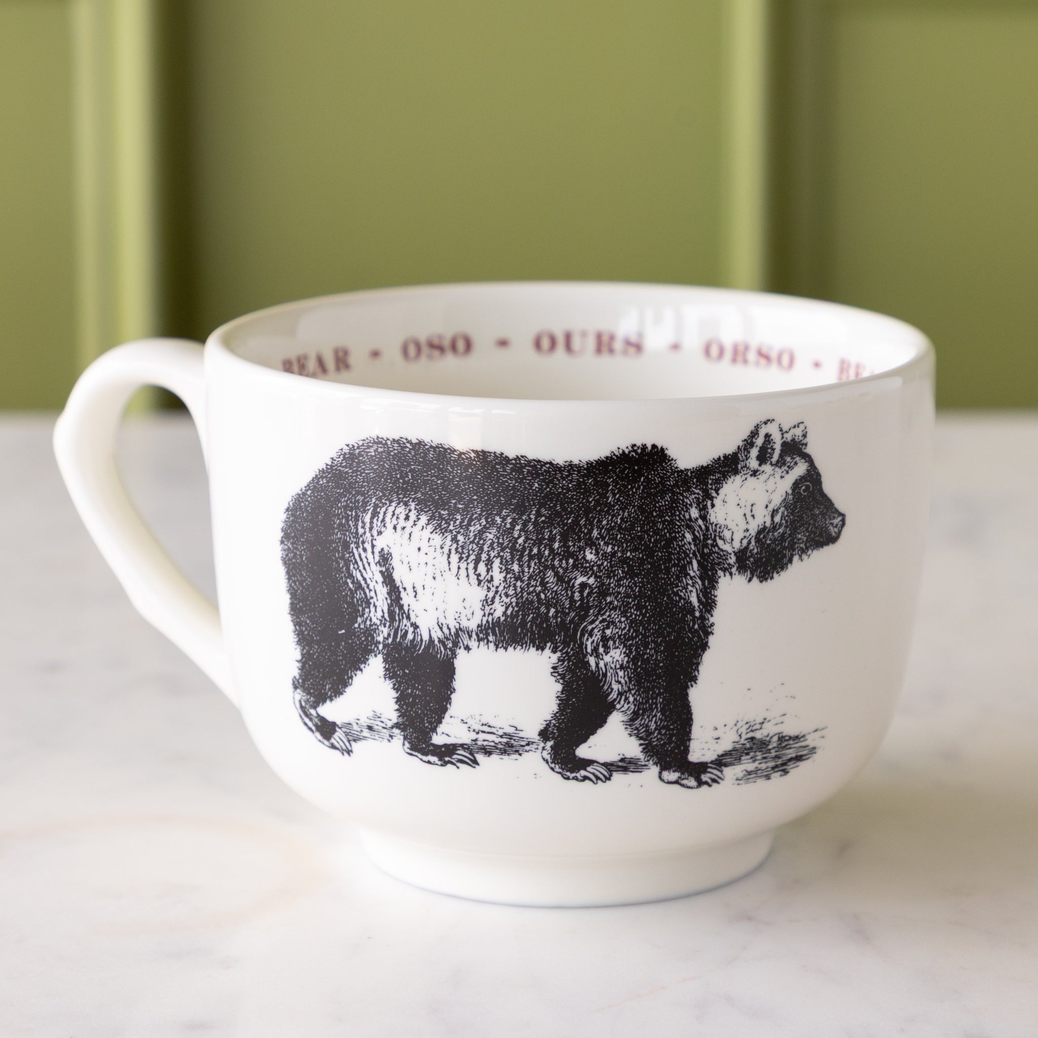 White mug with black bear illustration with "bear" in different languages in red text around the inside rim.