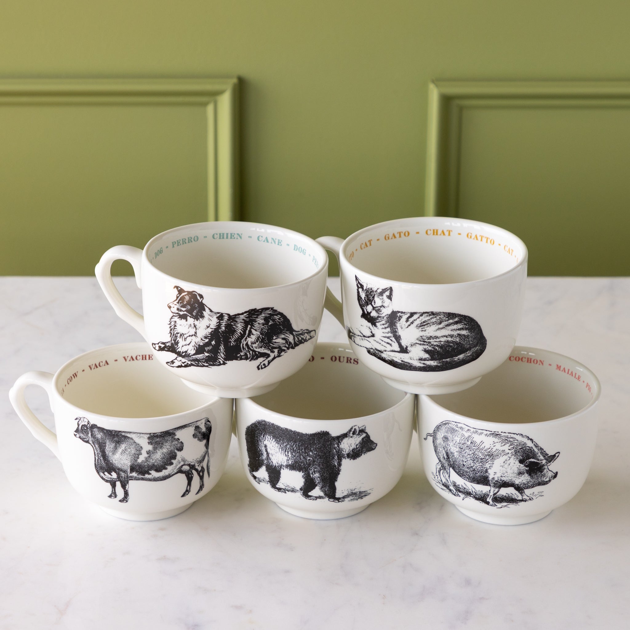 Set of five white mugs with black animal illustrations on a marble surface with a green wall background.