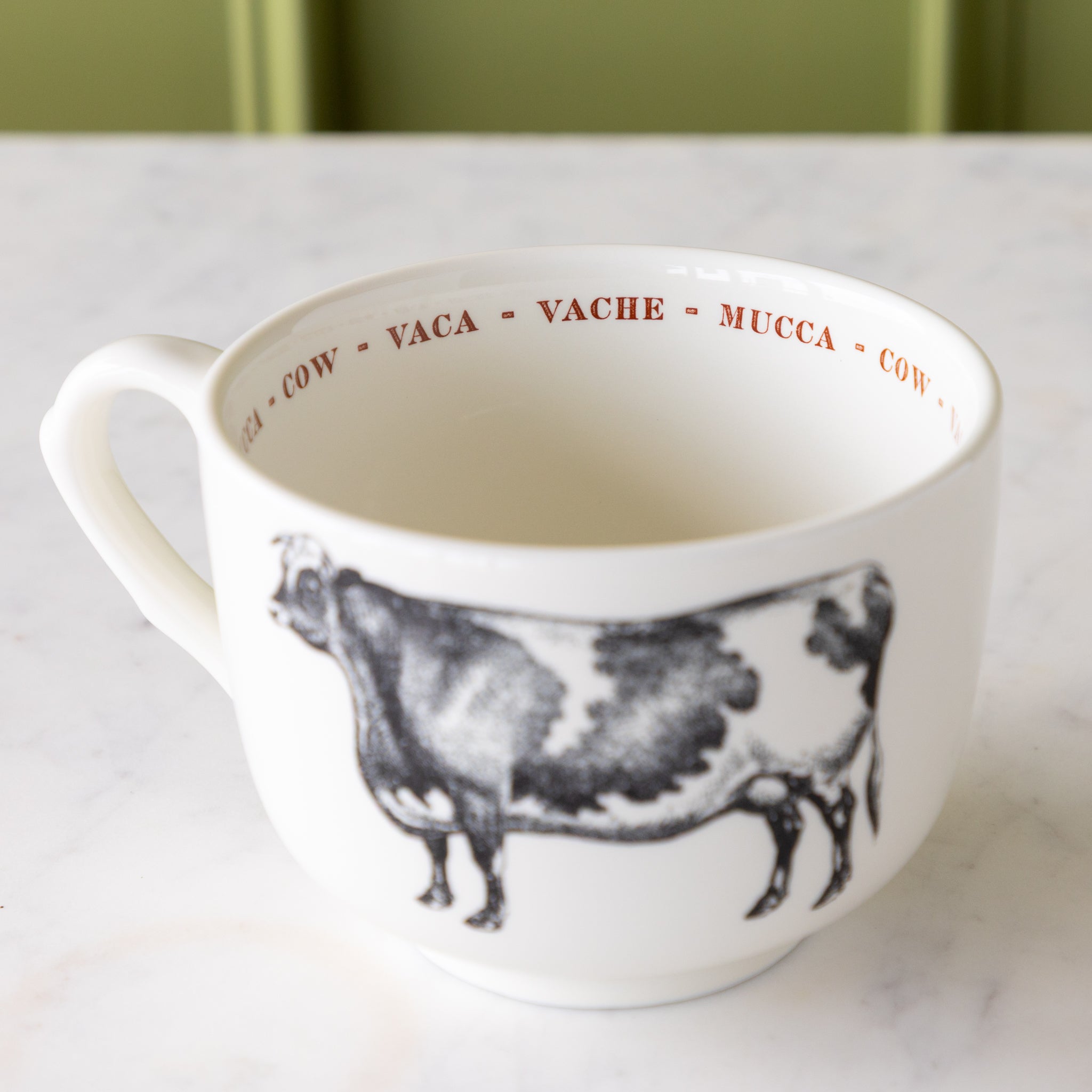 White mug with black cow illustration with "cow" in different languages in red text around the inside rim.