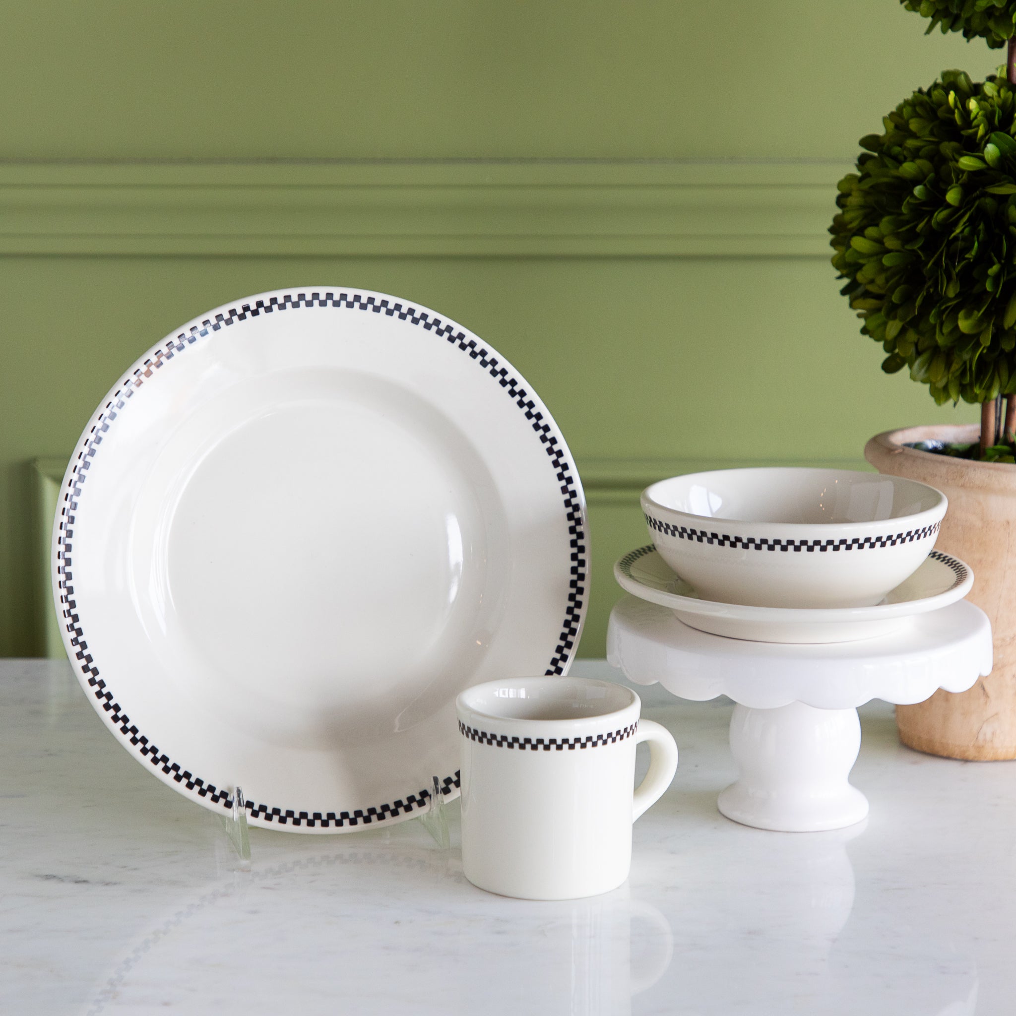 White dinnerware set with black checkered trim on a white surface with a green wall background.