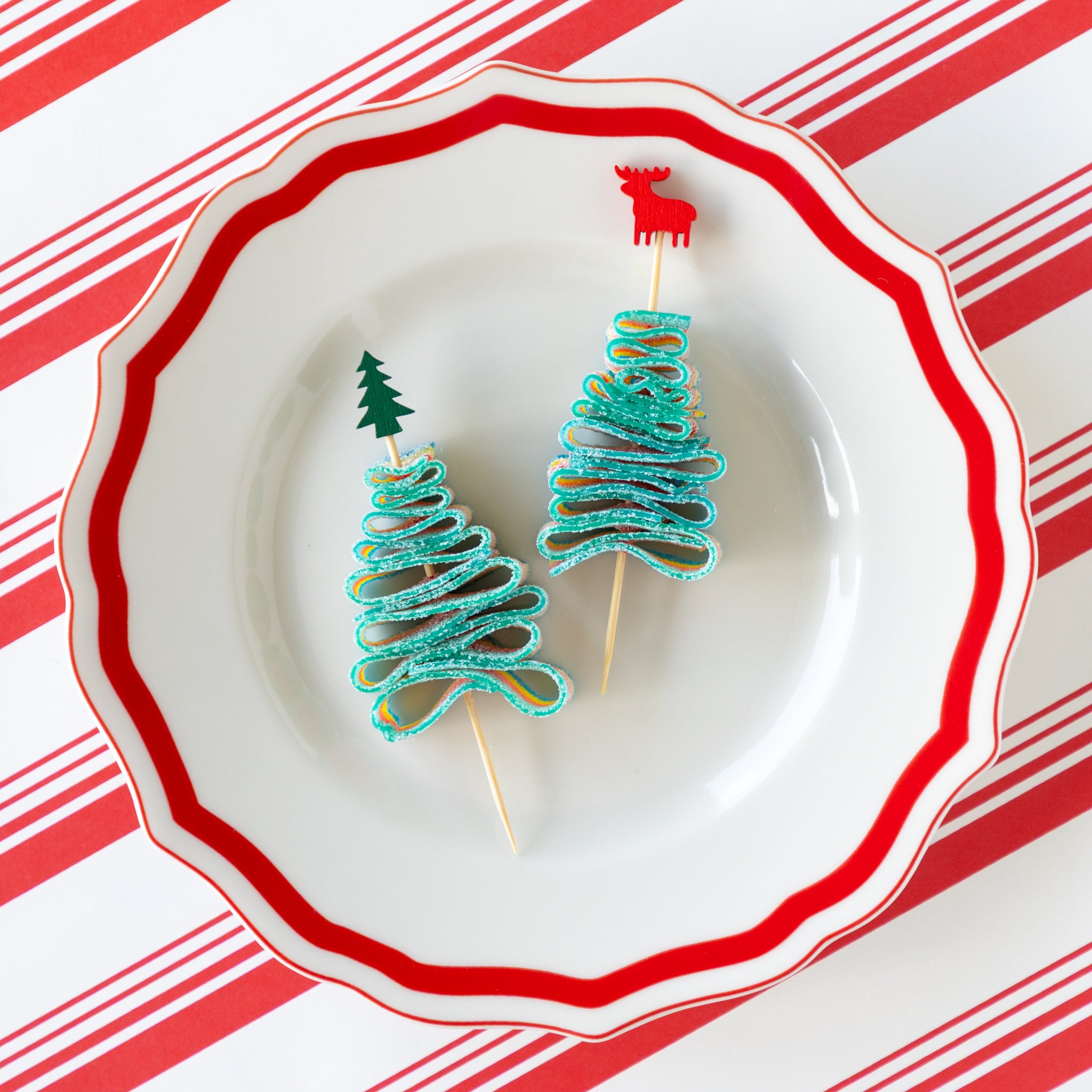 Deer and tree bamboo skewers piercing a candy rainbow rope folded in the shape of a tree, on a decorative plate and Peppermint Stripe Runner underneath.