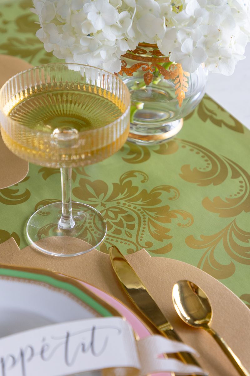 Table setting with gold cutlery, a glass of champagne, and a decorative green and gold floral paper table runner.