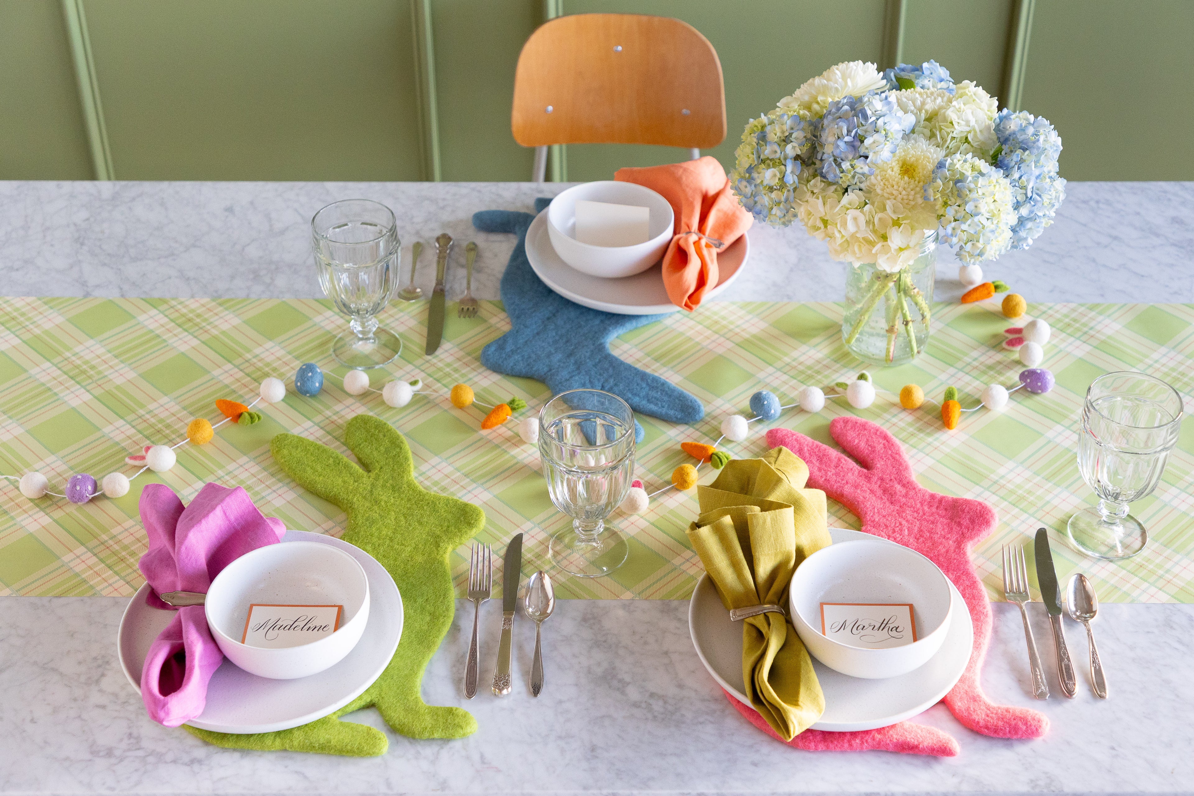 Easter-themed table setting with flowers, felt Spring Green, Pink & Cornflower Blue Easter Bunny Placemats, Easter Felt Garland and white dinnerware on the Green Plaid Runner.