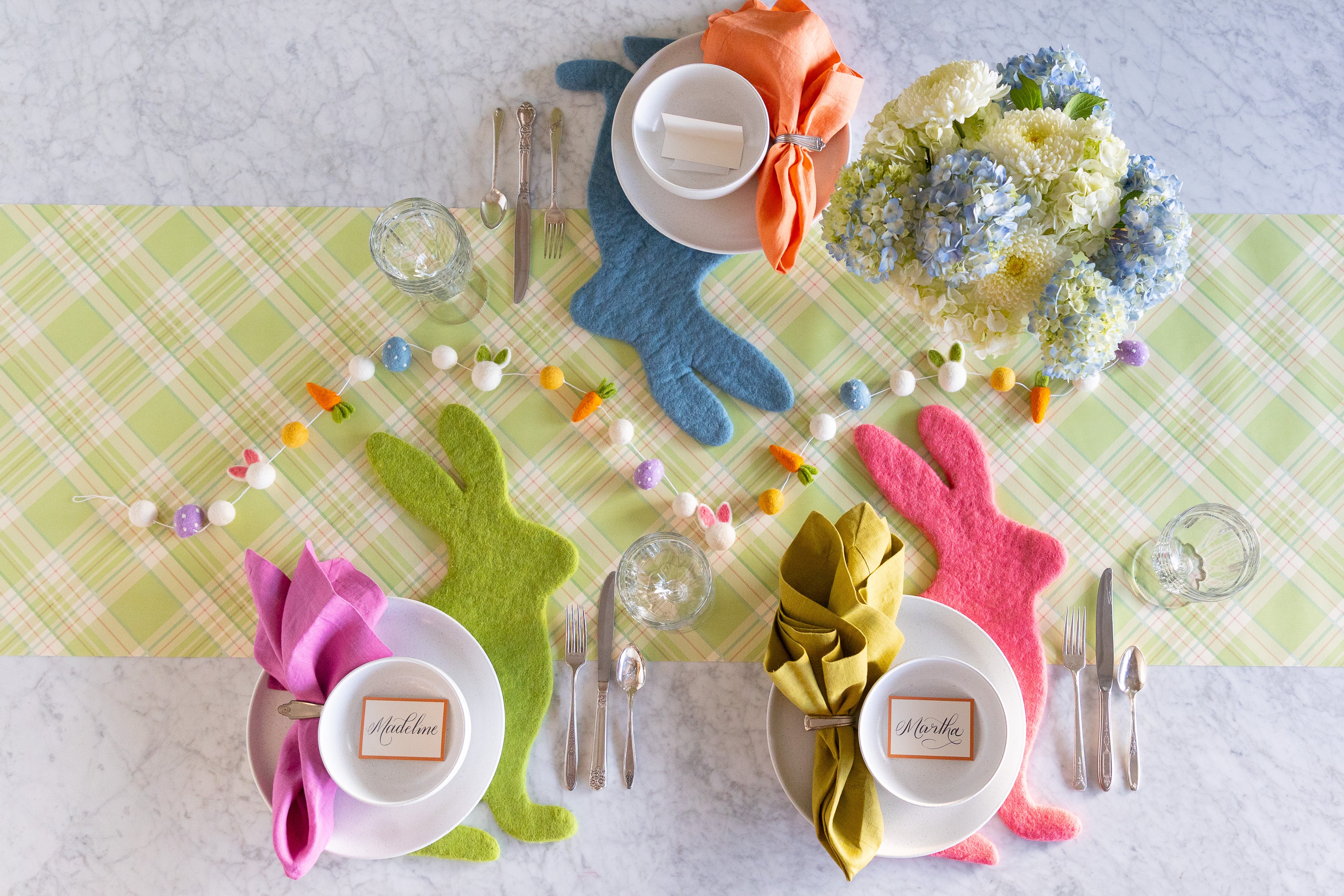 Easter themed table setting featuring flowers, Easter Felt Garland, Cornflower Blue, Spring Green and Pink Pom Felt Placemats on the Green Plaid Runner.