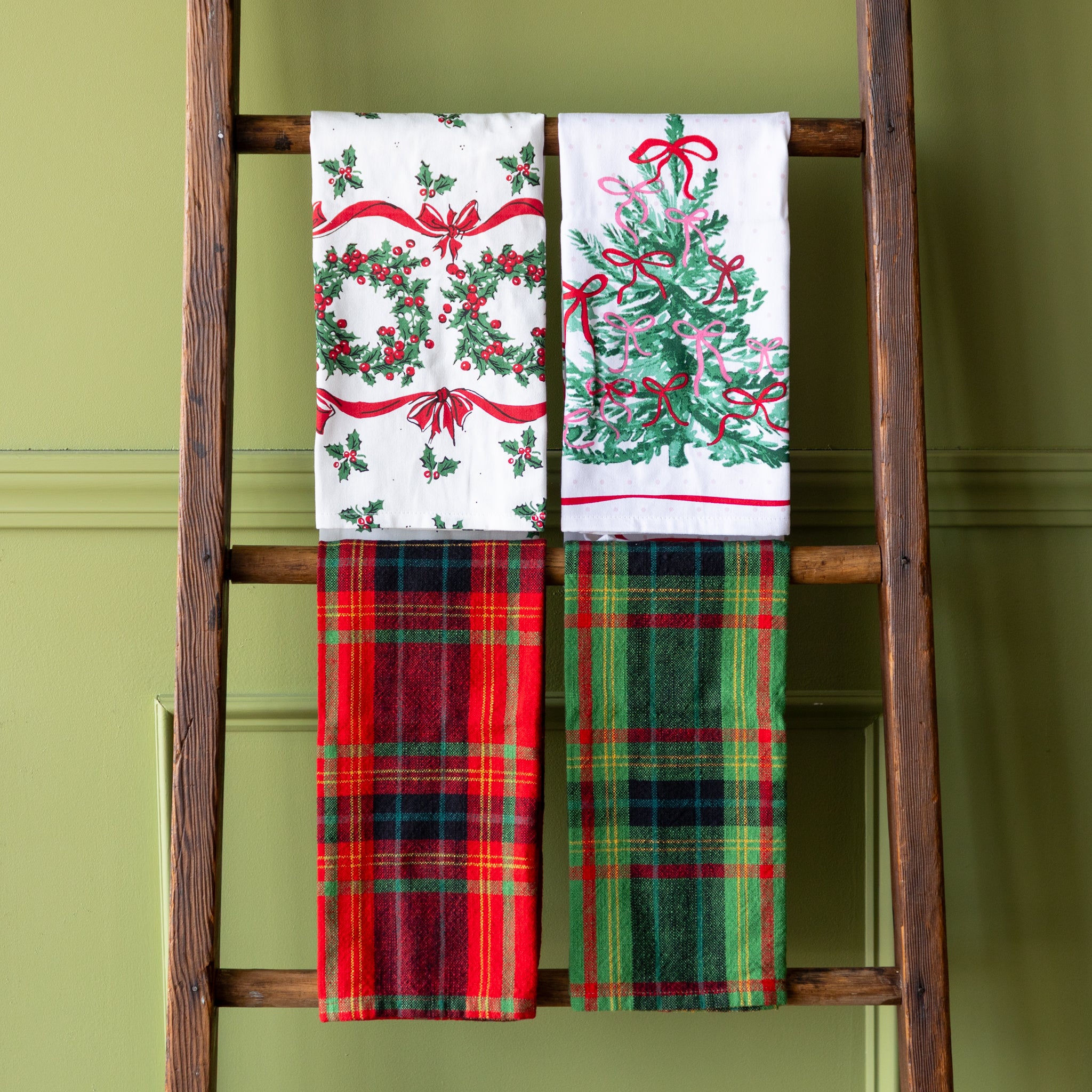 Four holiday tea towels hung on a wooden ladder against a green background.