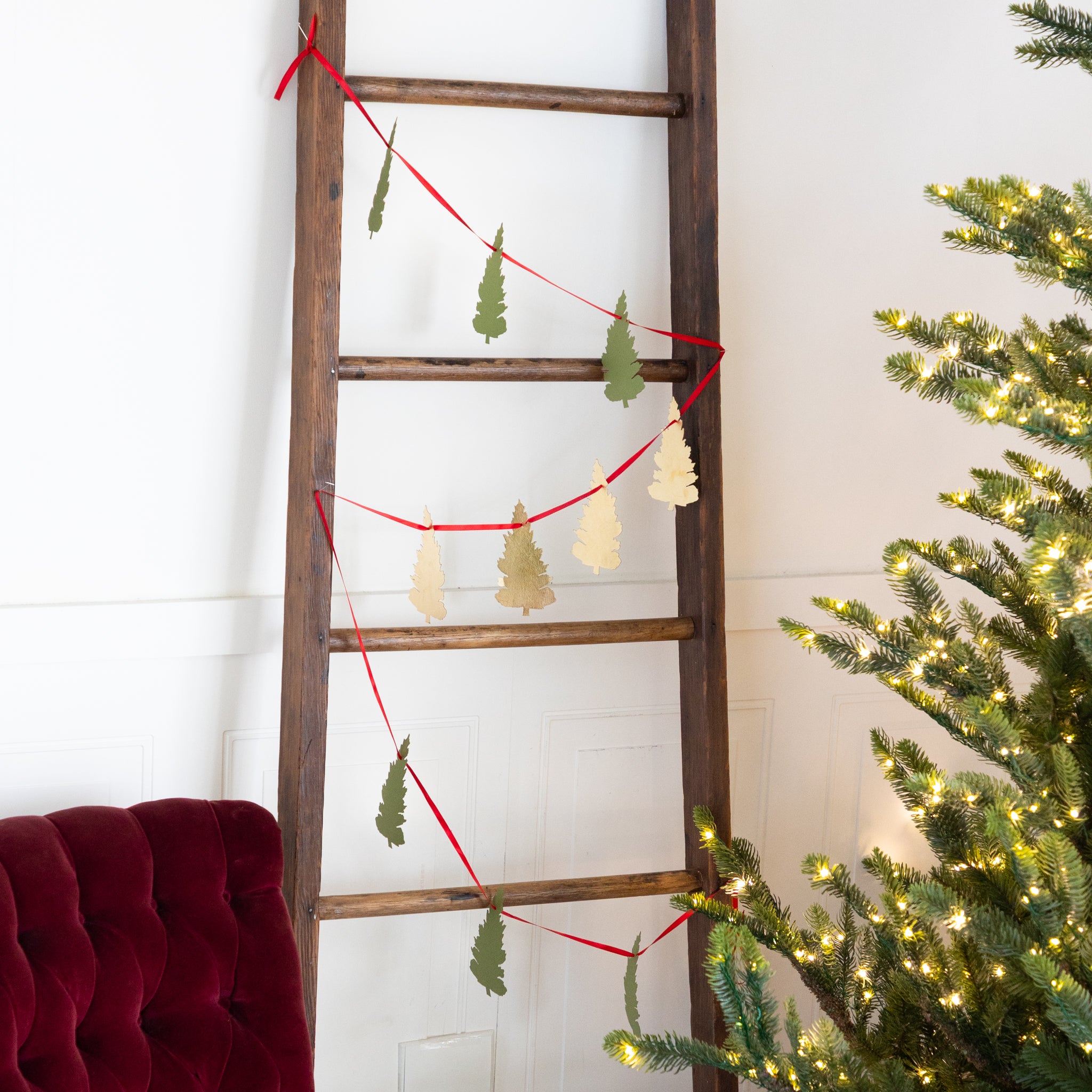 A garland of shaped evergreen trees tied to a wooden ladder next to a lit Christmas tree.