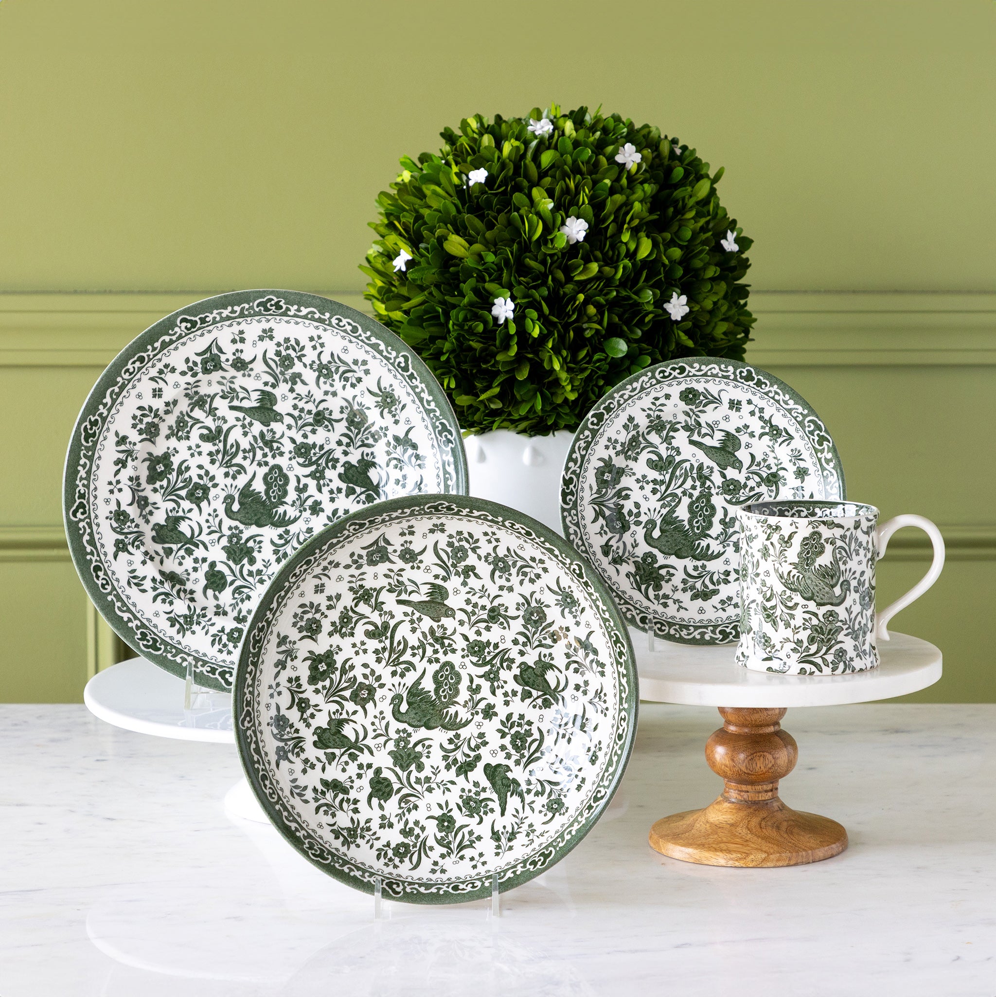 Burleigh Green Regal Peacock Dinnerware displayed on a table with topiary behind it.