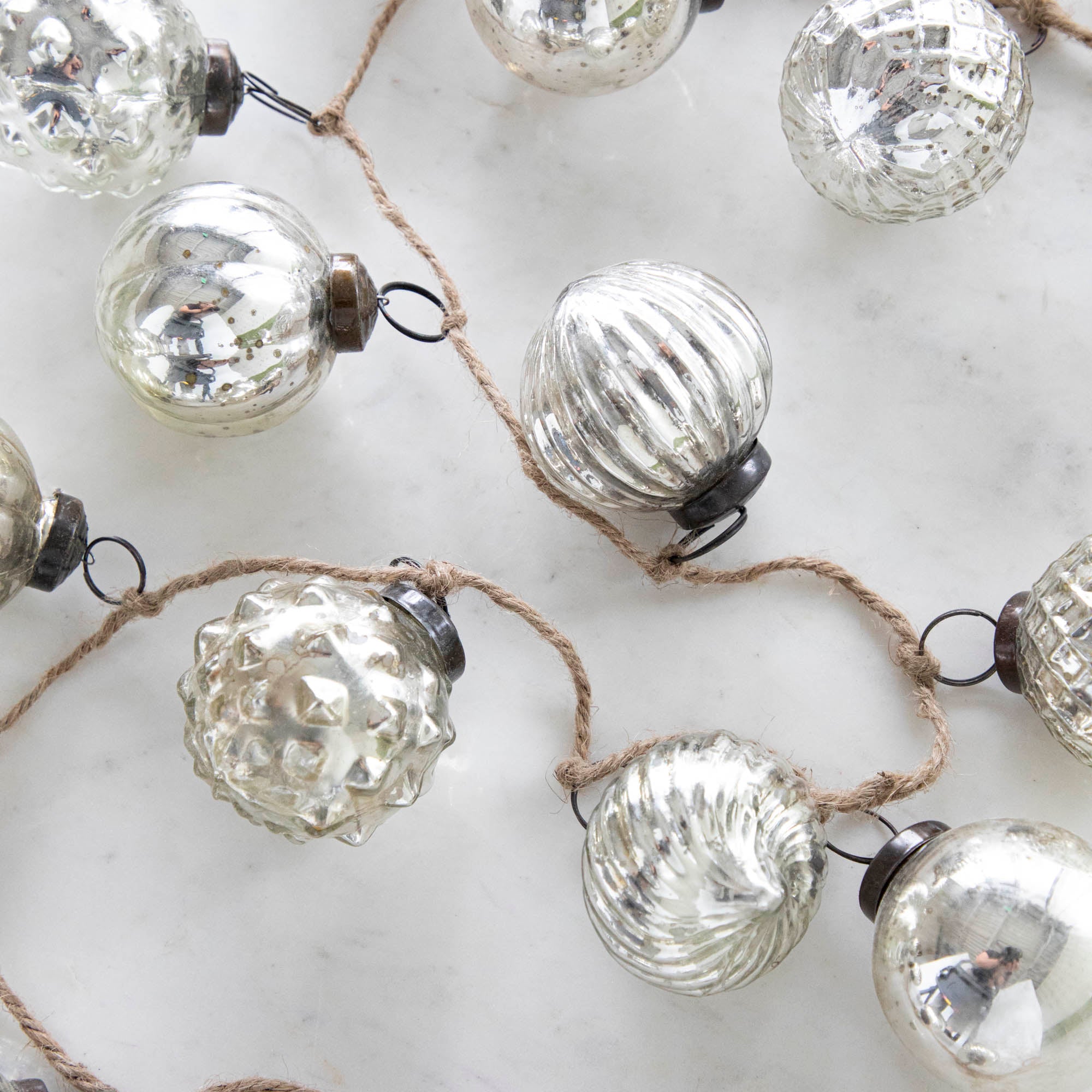Silver Embossed Mercury Glass Ornament Garland with a jute cord on a marble table.