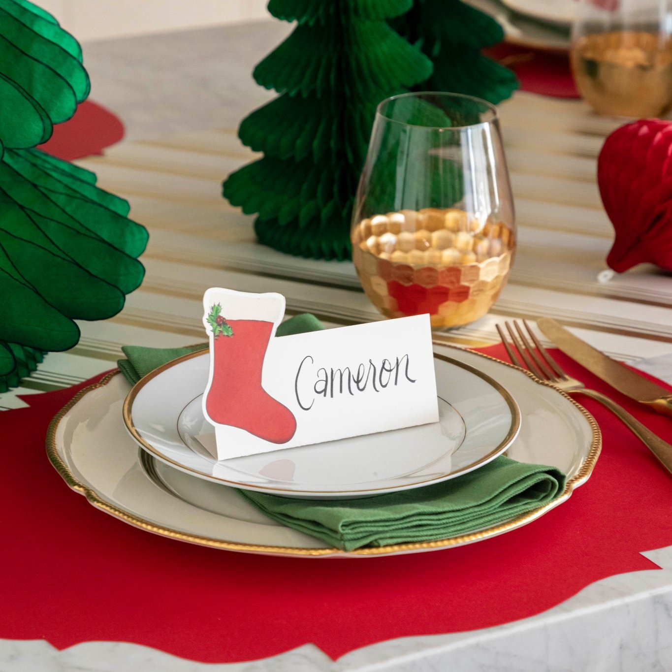 An elegant Christmas themed table setting with the Die-cut Red French Frame Placemat under gold rimmed plates, a green napkin, and a Stocking Place Card with the name "Cameron" written on it.