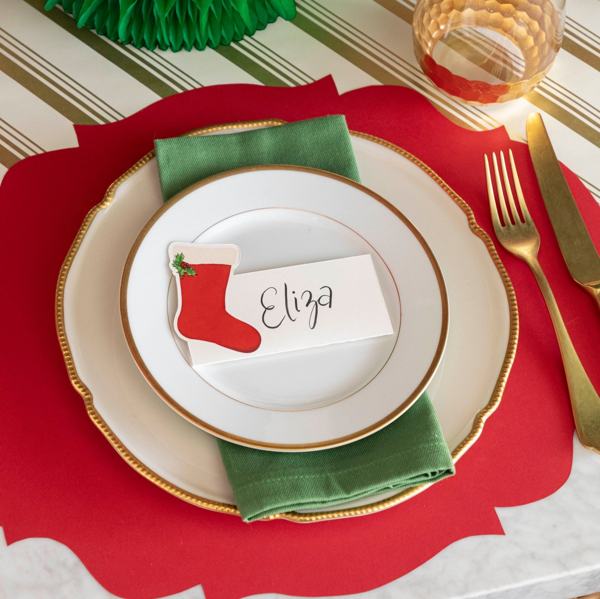 An elegant table setting with the Die-cut Red French Frame Placemat on the Gold Peppermint Stripe Runner, with gold rimmed plates and a a Stocking Place Card with the name "Eliza" written on it, on top.