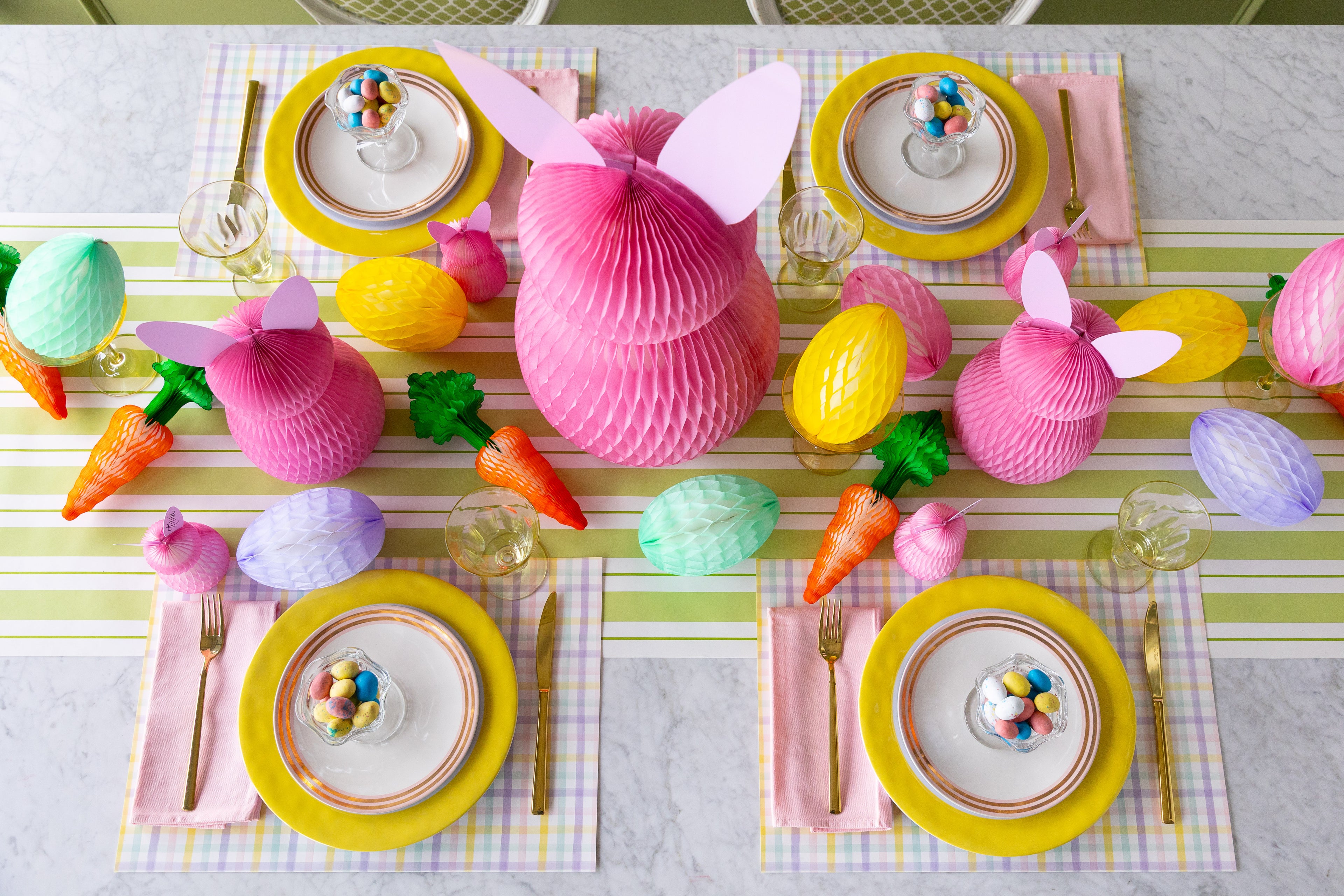 Easter-themed table setting with honeycomb bunnies, eggs and carrots decorations, Spring Plaid Placemats, colorful plates, pink napkins and gold flatware on the Green Awning Stripe Runner.