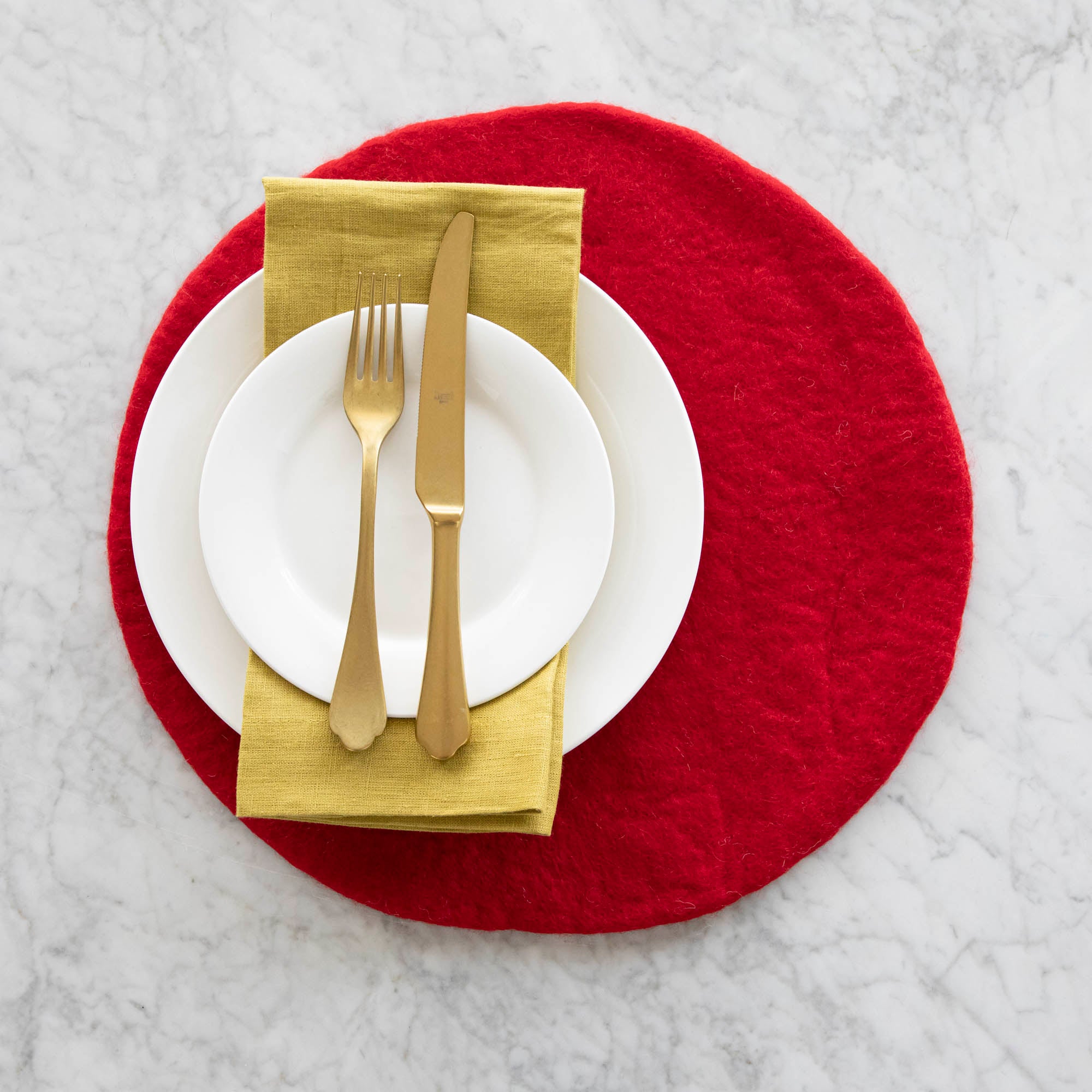 A place setting with the Red Felt Placemat, white dinner plates with a gold napkin in between and a gold fork and knife atop, on a marble table.