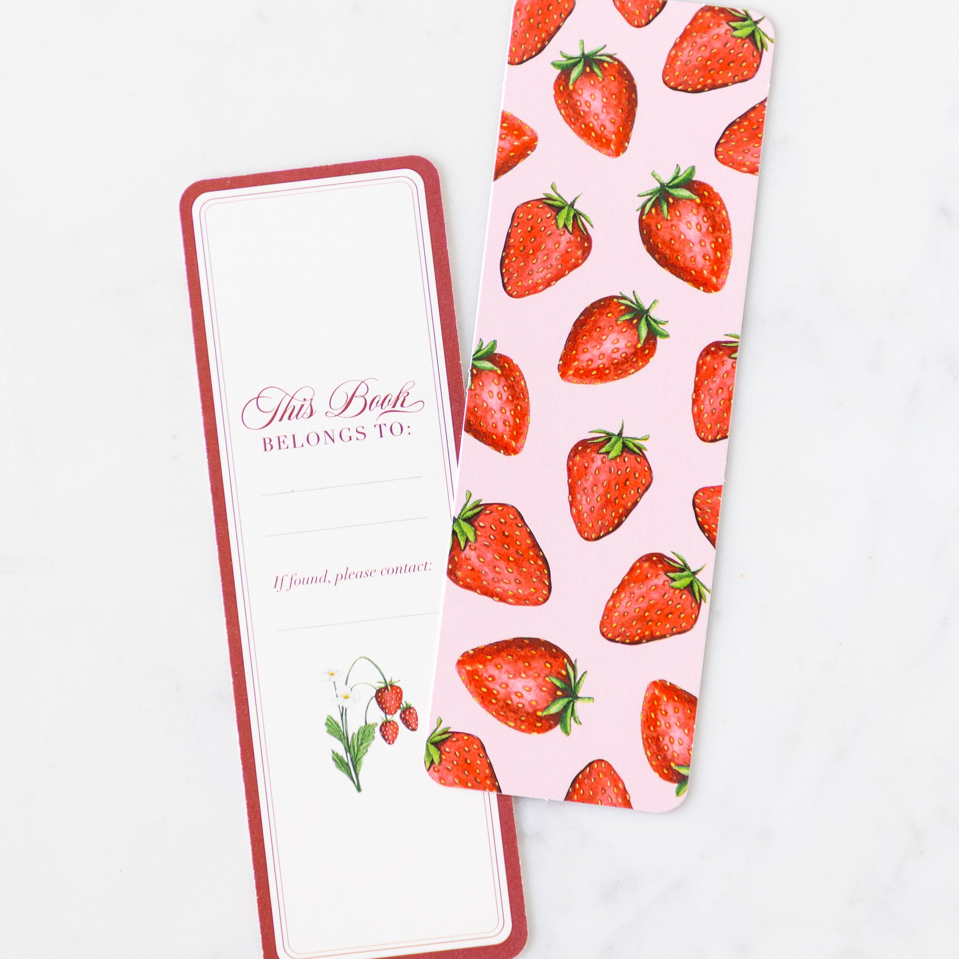 Two Berries & Cherries Bookmarks on a marble table––the front features a strawberry design with a pink background while the back features a red border, "This Book Belongs To:", a place to write a name and a strawberry plant.