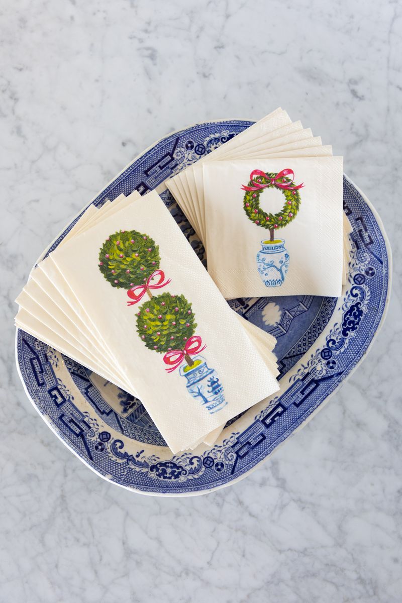 Two fanned stacks of napkins, guest size and cocktail size, featuring a holiday topiary design on a blue and white platter on a white marble surface.