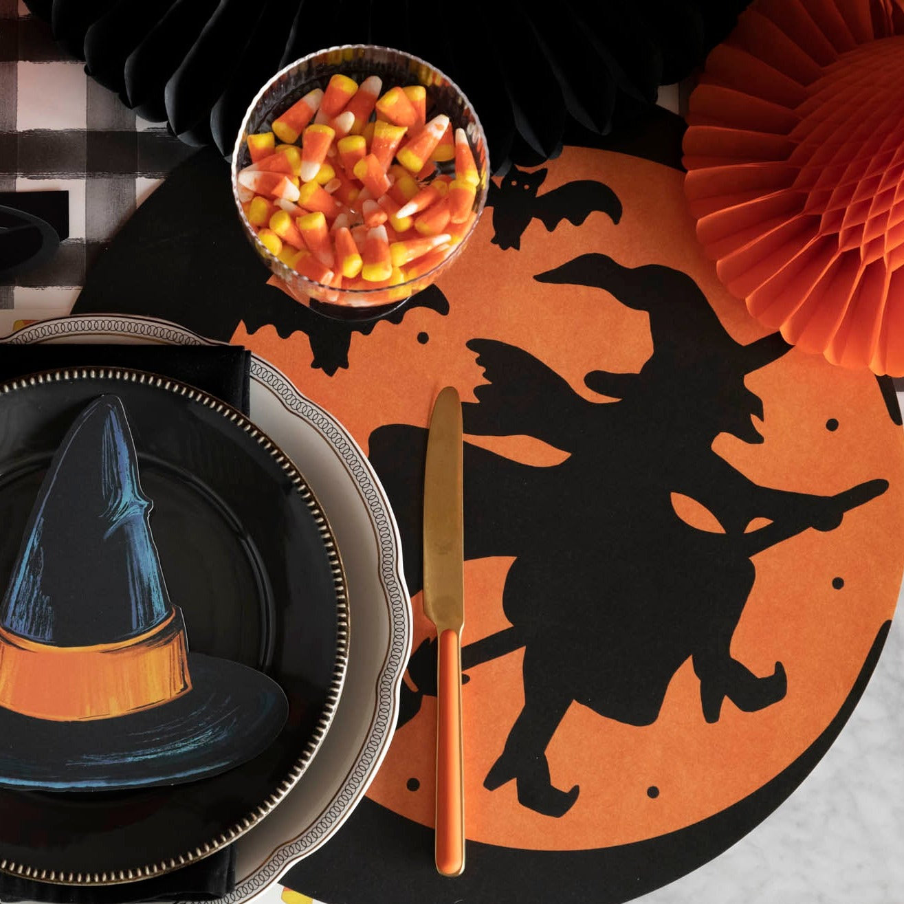 A Halloween-themed table setup featuring a die-cut witch placemat, a plate with the Witch H Table Accent, a bowl of candy corn, and the Die-cut Wicked Witch Placemat.