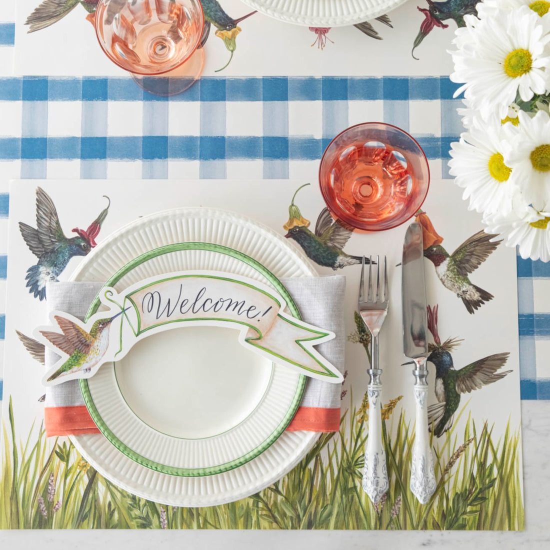Top-down view of a springtime place setting featuring a Hummingbird Banner Table Accent with &quot;Welcome!&quot; written on it resting on the plate.