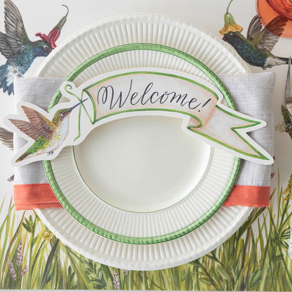Close-up of a springtime place setting featuring a Hummingbird Banner Table Accent with &quot;Welcome!&quot; written on it resting on the plate.
