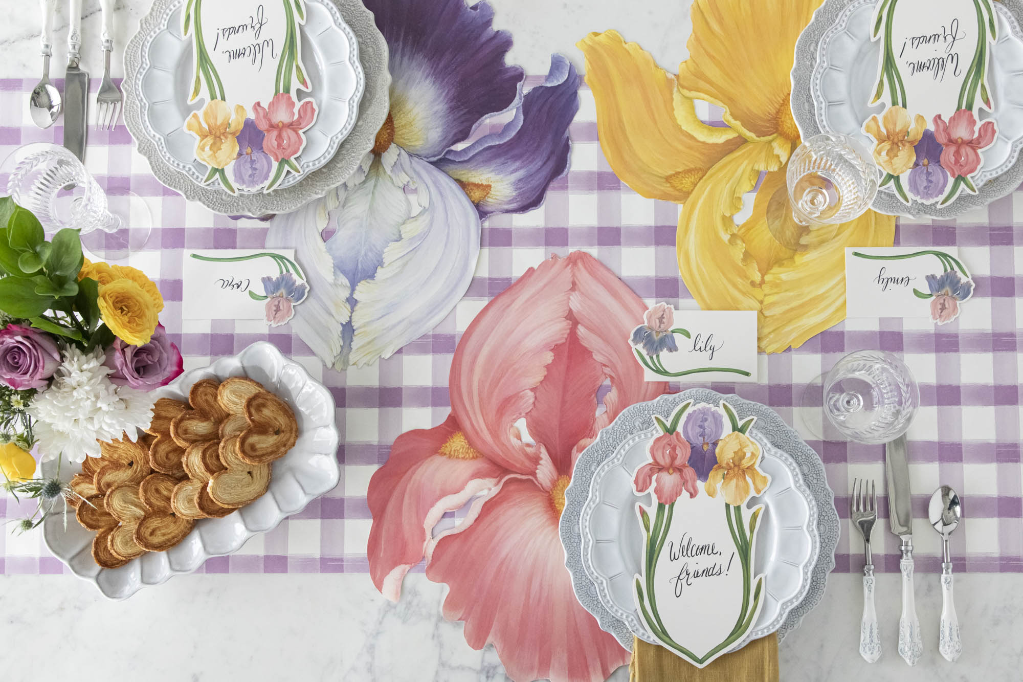 Top-down view of an elegant floral table setting featuring an Iris Table Card with "Welcome, friends!" written on it in beautiful script resting on each plate.