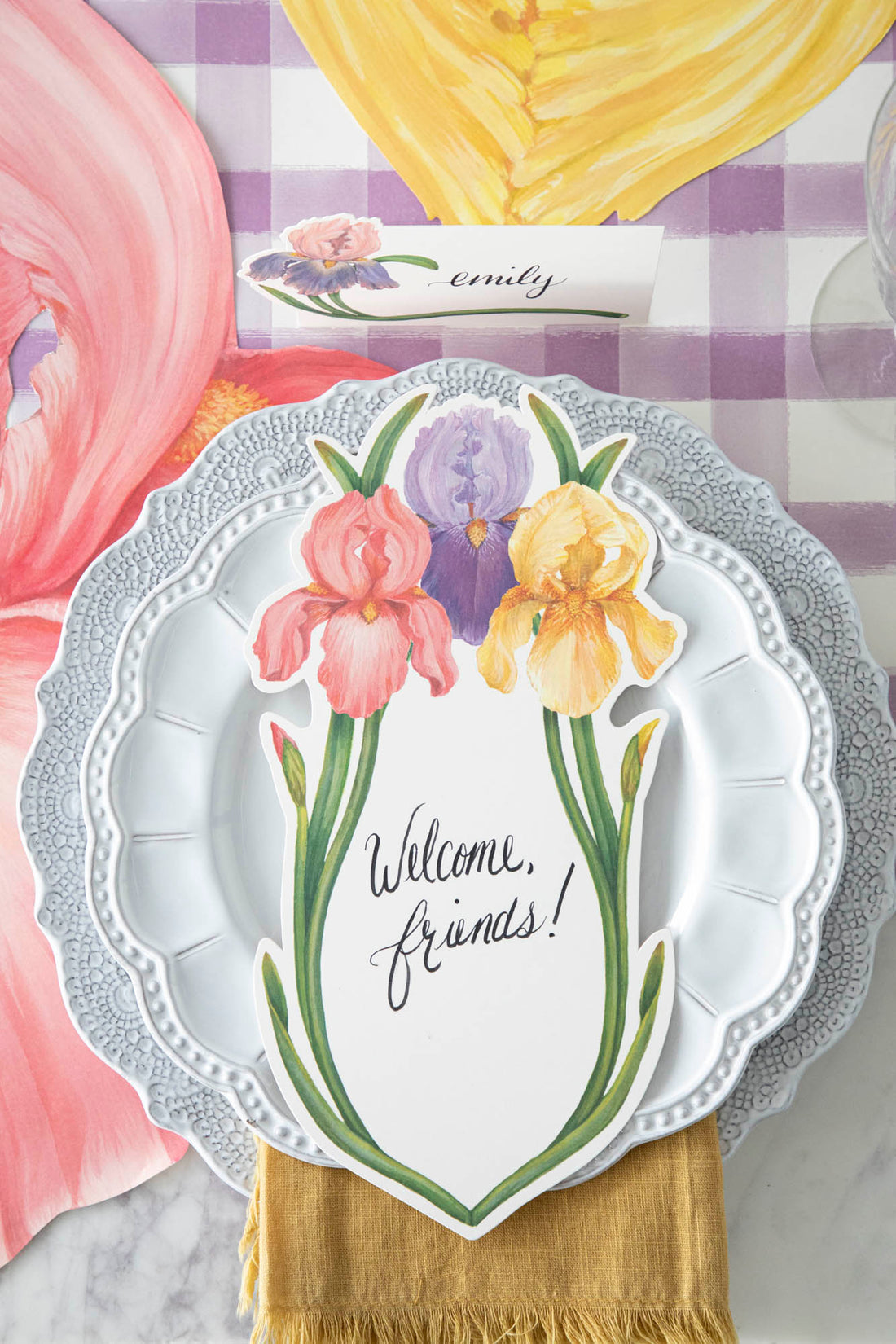 Top-down view of an elegant floral place setting featuring an Iris Table Card with &quot;Welcome, friends!&quot; written on it in beautiful script resting on the plate.