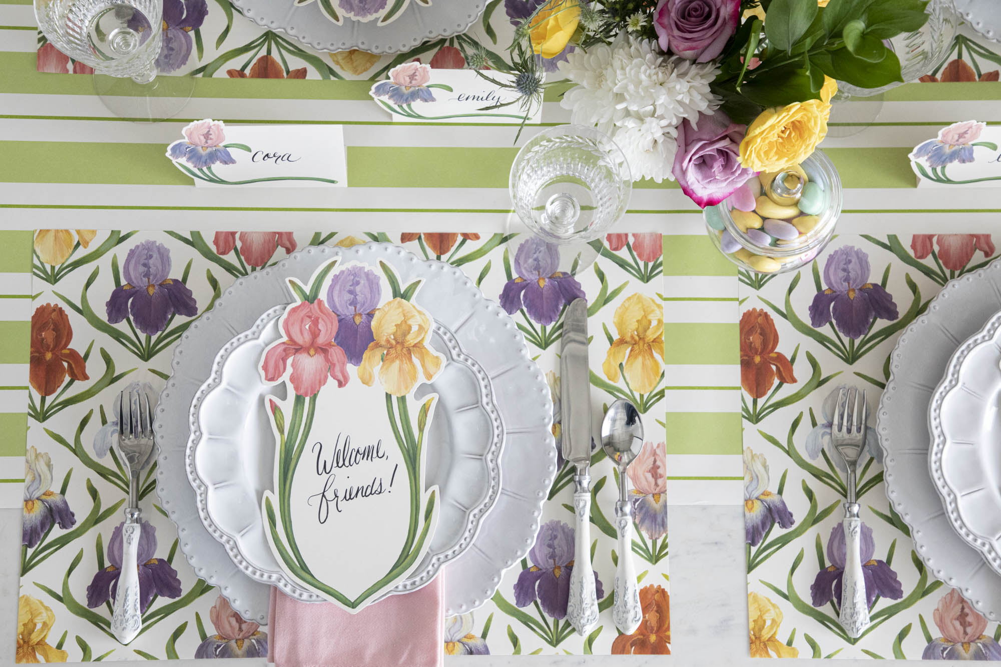 Top-down view of an elegant floral place setting featuring an Iris Table Card with &quot;Welcome, friends!&quot; written on it in beautiful script resting on the plate.