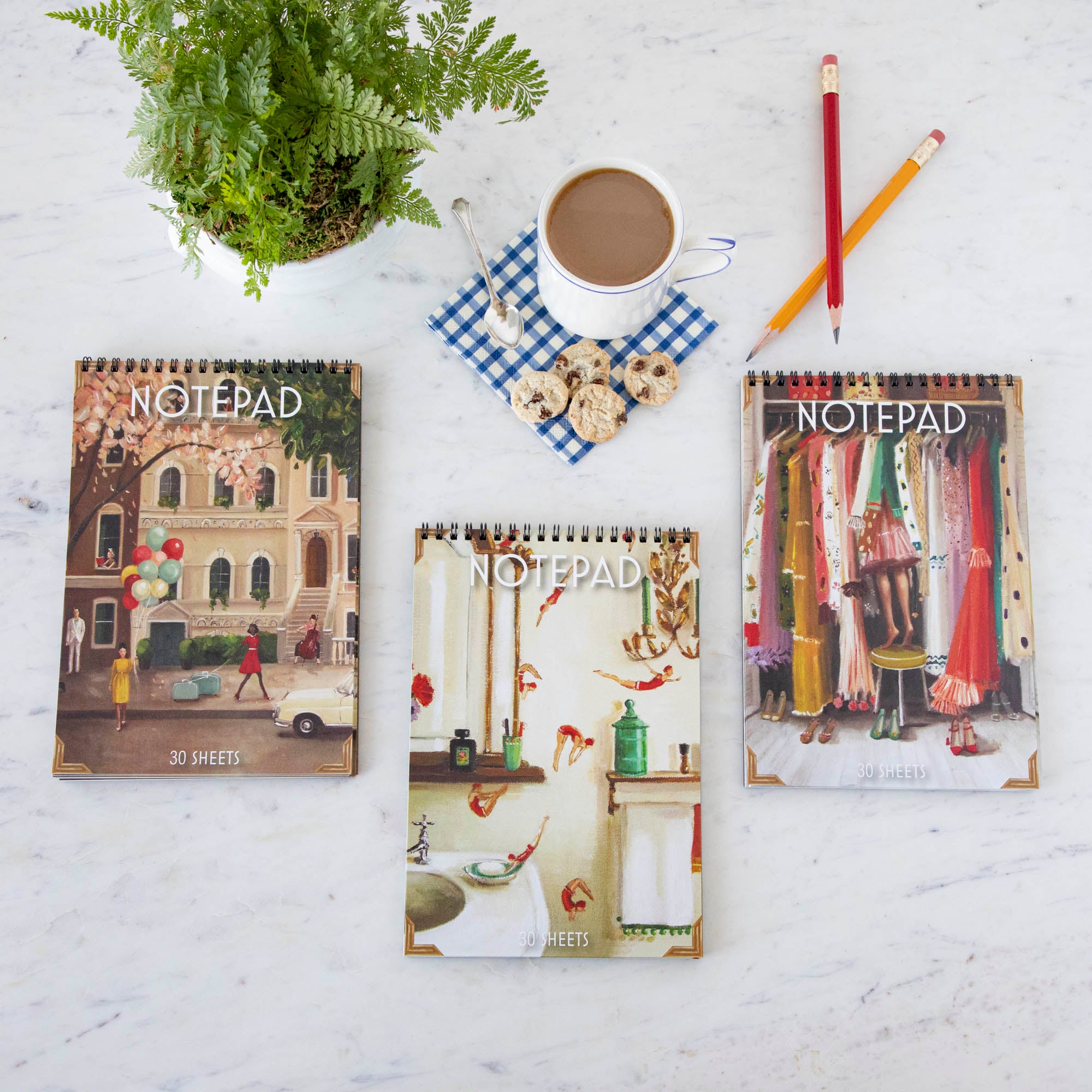 Three different Janet Hill Notepads resting on a white table with pencils and a cup of coffee.