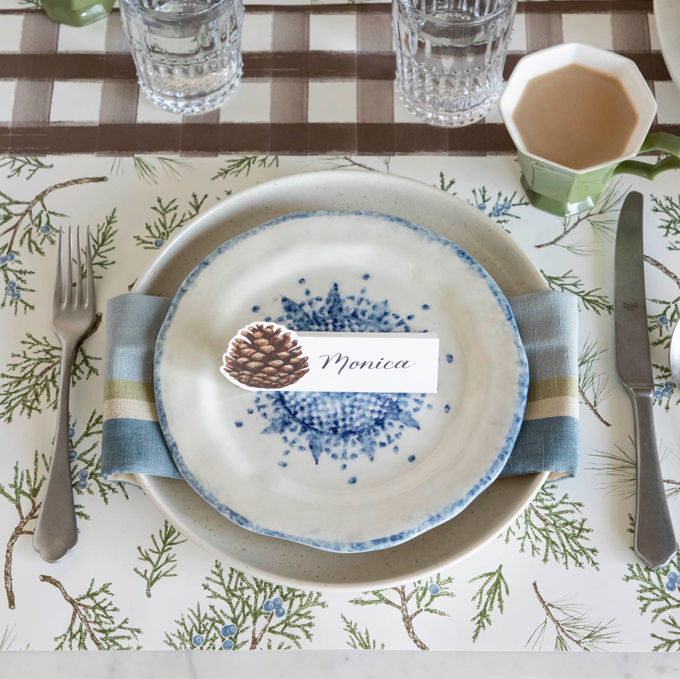 Elegant table setting with the Juniper Sprigs Placemat, a napkin in between two plates and a Pinecone Place Card with the name "Monica" written on it, on top.
