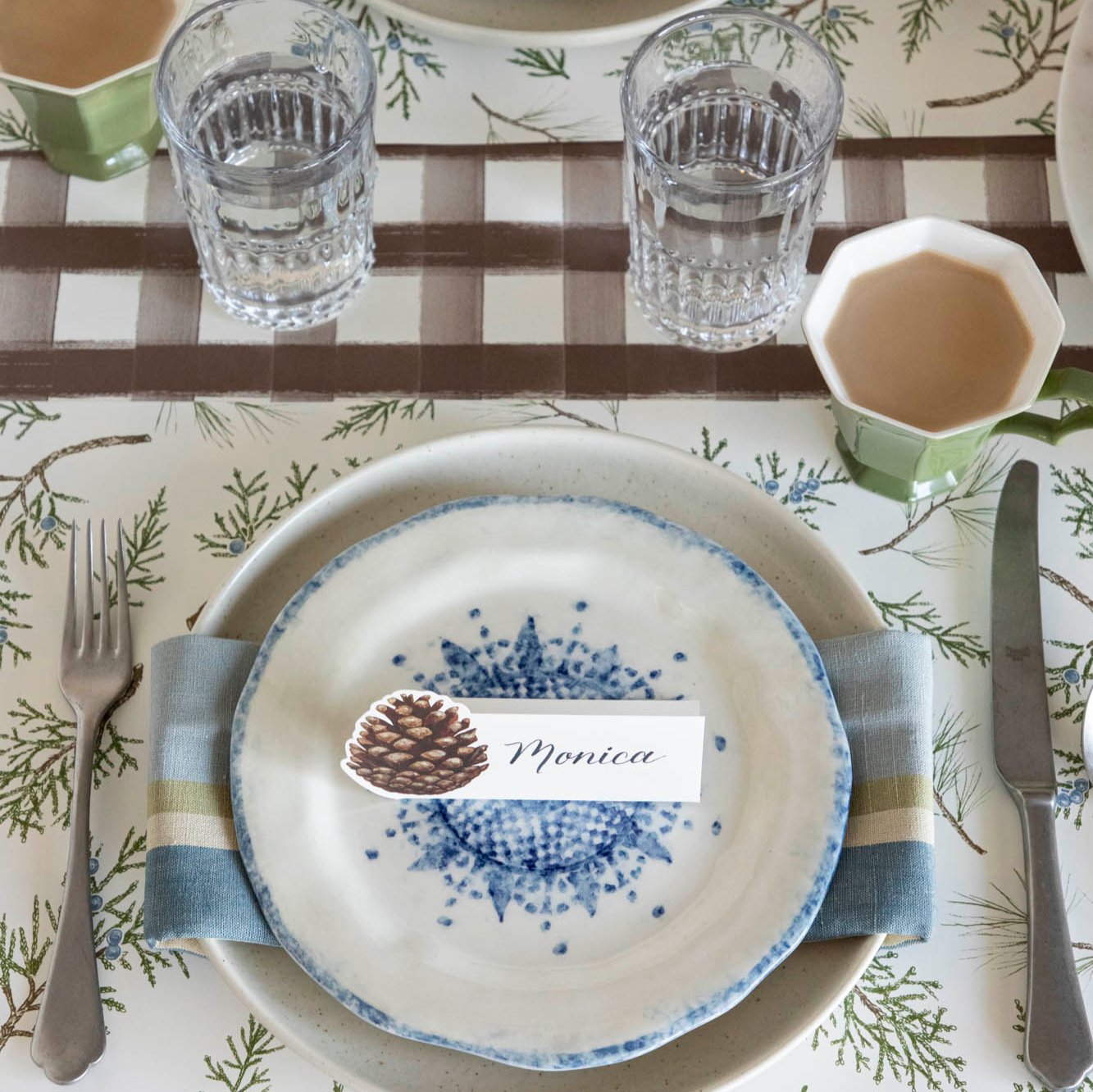 An elegant table setting with the Brown Painted Check runner underneath a Juniper Sprigs Placemat and dinnerware with a Pinecone Place Card with "Monica" written on it, on top.