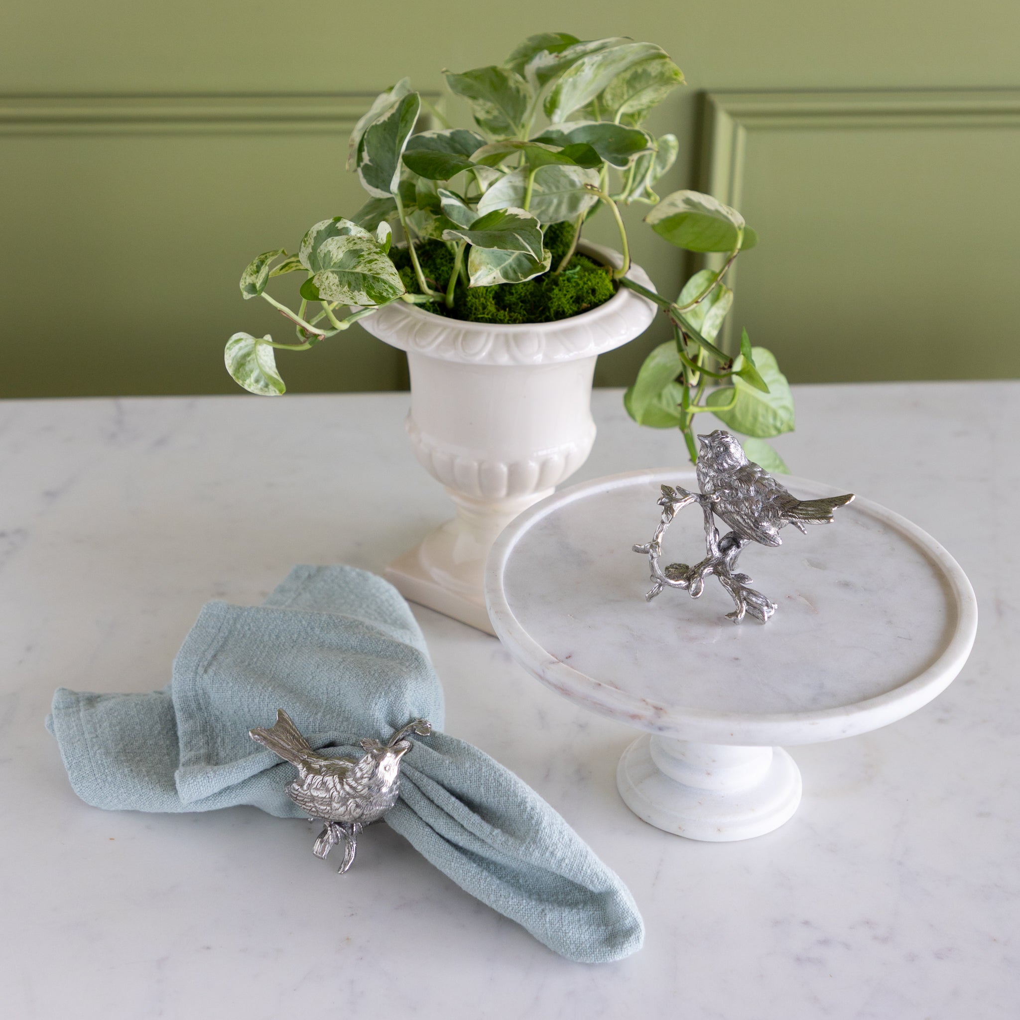 Decorative silver bird napkin ring on a light blue napkin and white pedestal with a plant in the background.
