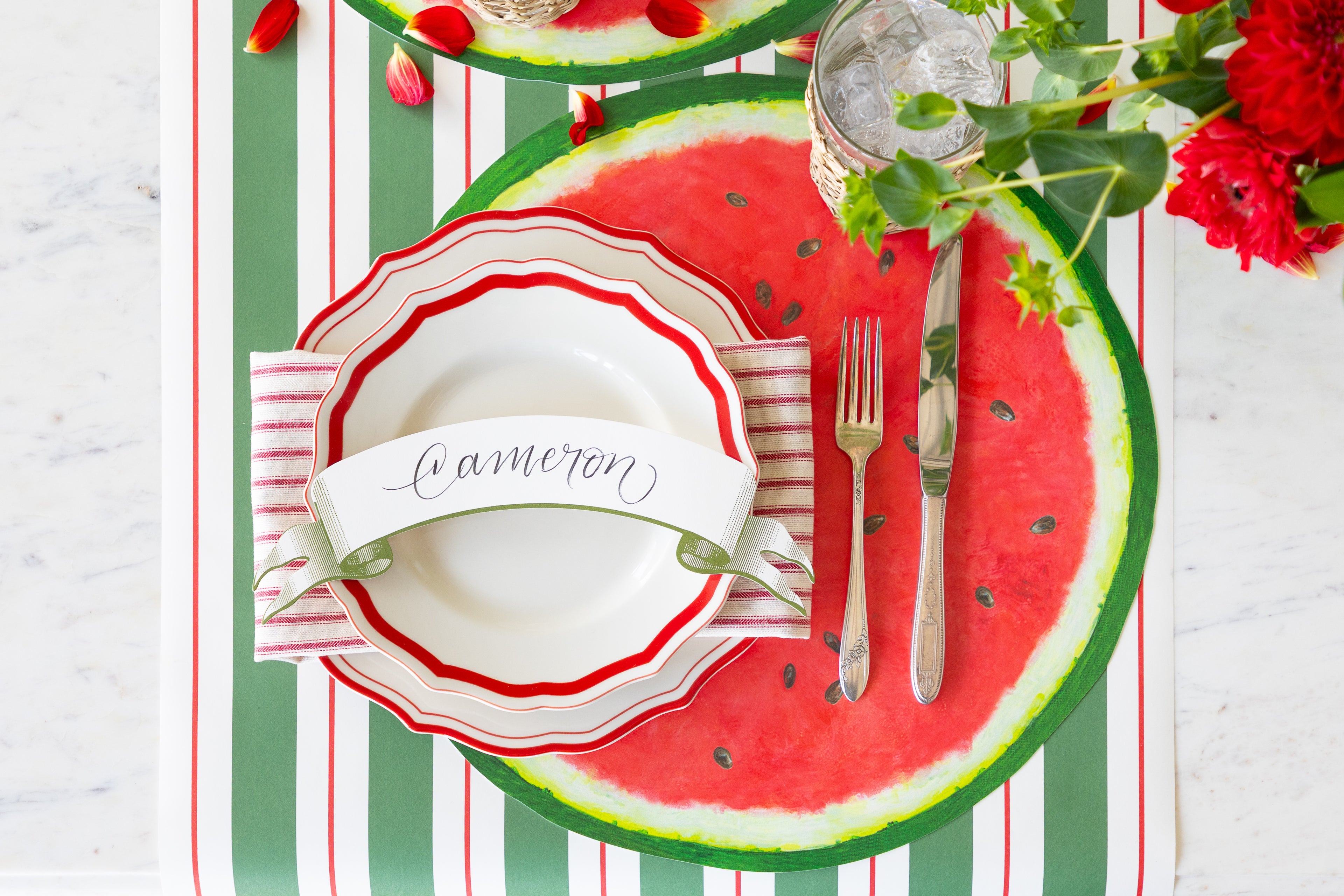 Table setting with watermelon-themed plates and cutlery on a striped tablecloth.