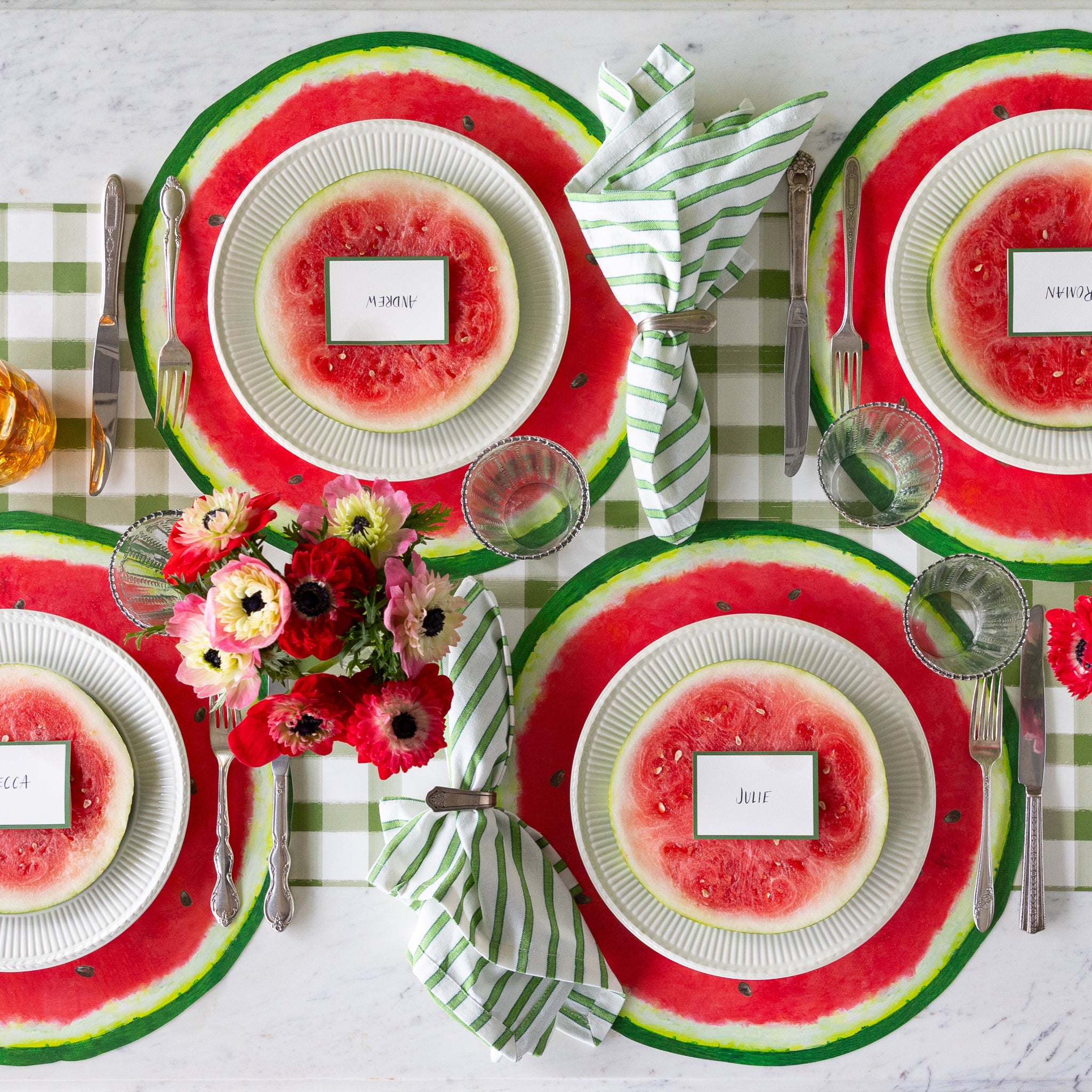 Table setting with Die-cut Watermelon Placemats, plates with watermelon slices and Dark Green Frame Place Cards atop, green striped napkins, glasses, silverware and a floral centerpiece on the Moss Green Painted Check Runner.