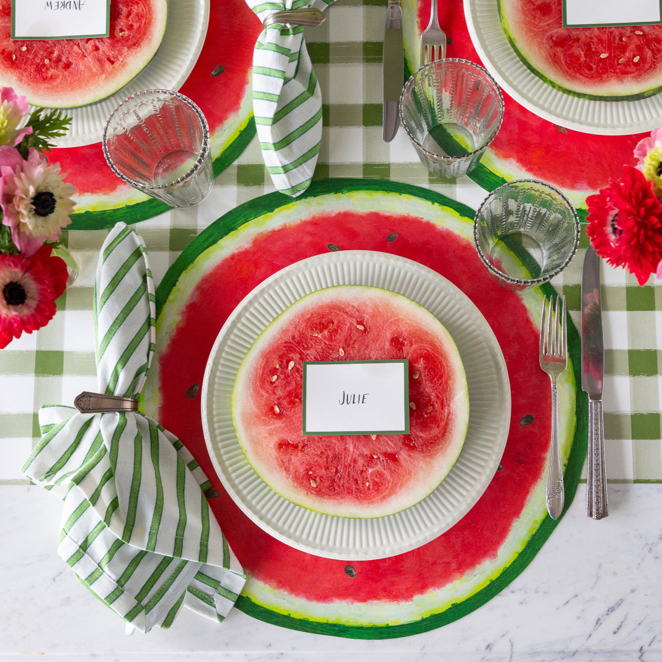 Table setting with Die-cut Watermelon Placemats, plates with watermelon slices and Dark Green Frame Place Cards atop, green striped napkins, glasses, silverware and a floral centerpiece on the Moss Green Painted Check Runner.