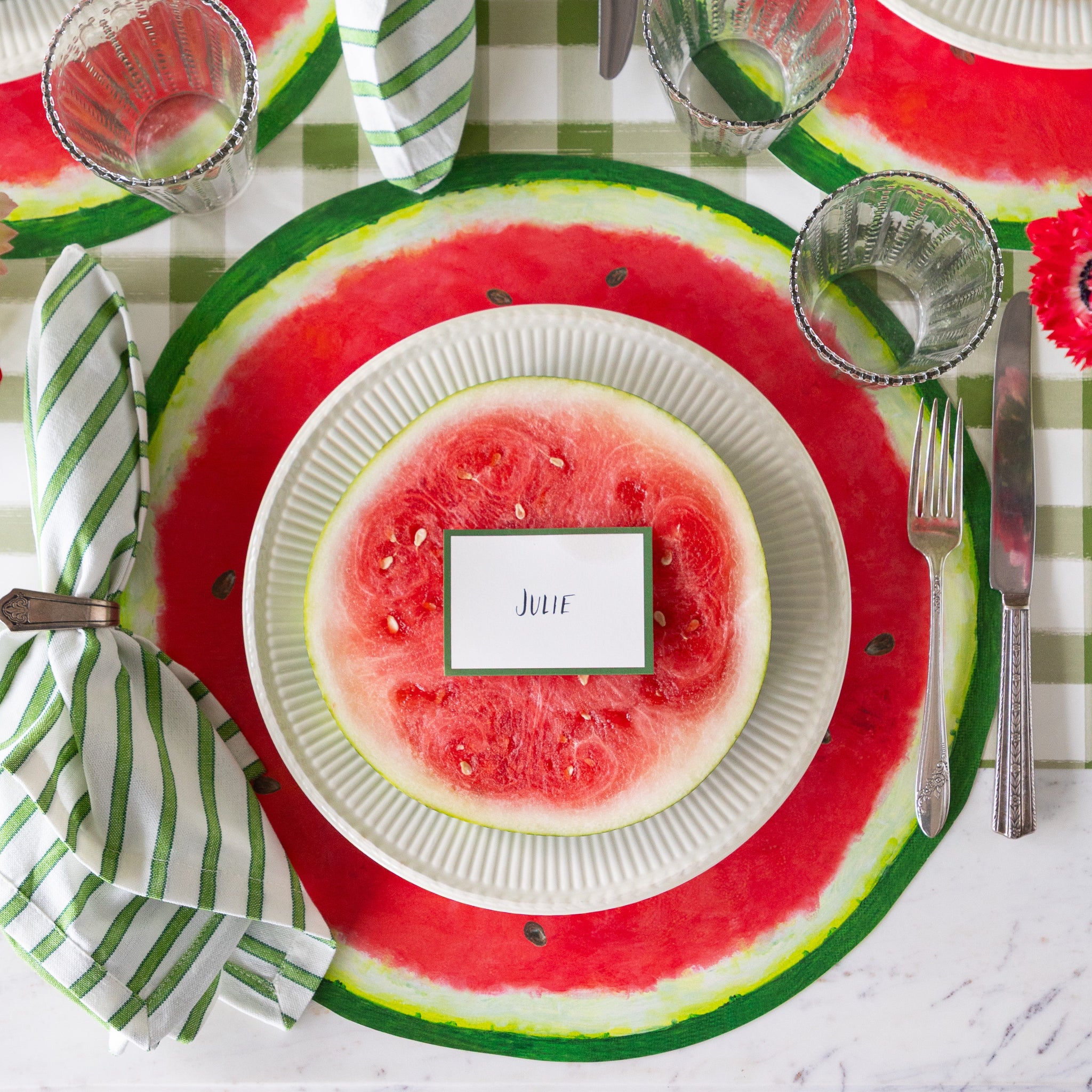 Place setting with the Die-cut Watermelon Placemat, plate with a watermelon slice and Dark Green Frame Place Card atop, silverware and glasses on the Moss Painted Check Runner.