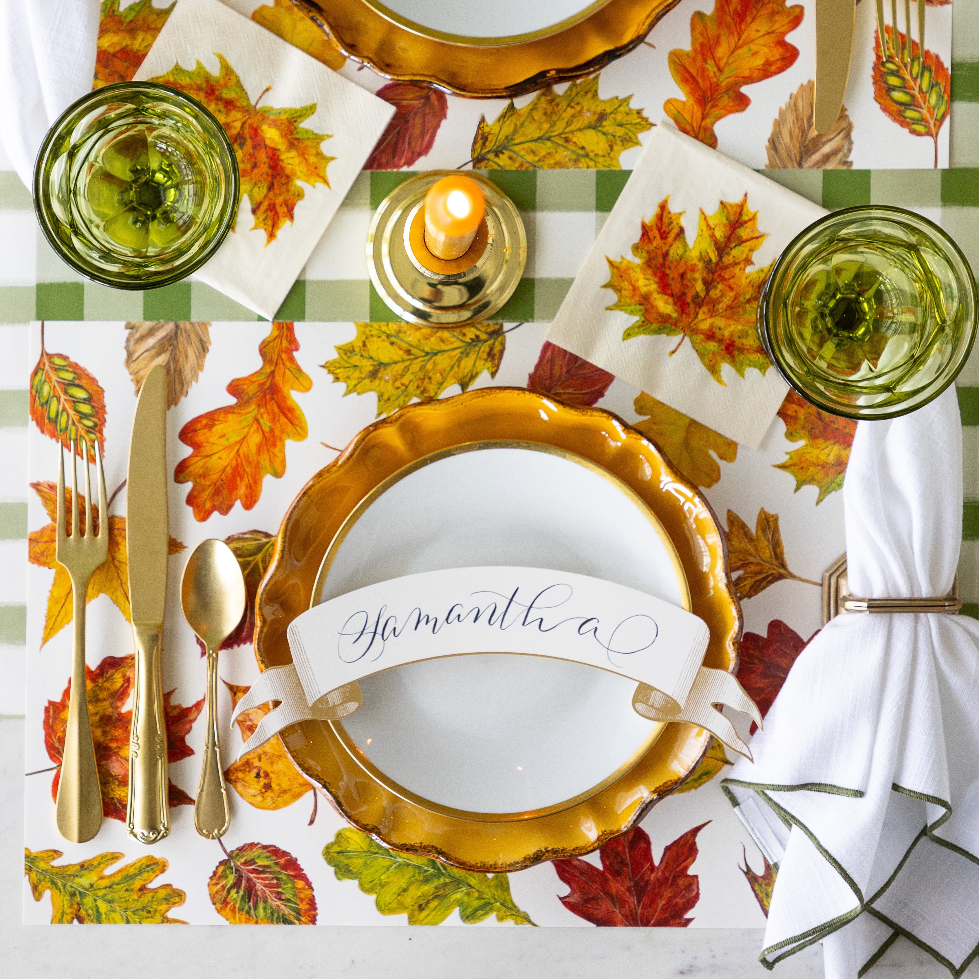 A place setting featuring the Fall Foliage Placemat under Mepra gold flatware, dinnerware with the Classic Gold Banner Table Accent reading "Samantha", atop, a napkin in a holder, the Fall Foliage Cocktail Napkin under a glass and Moss Green Painted Check Runner underneath.