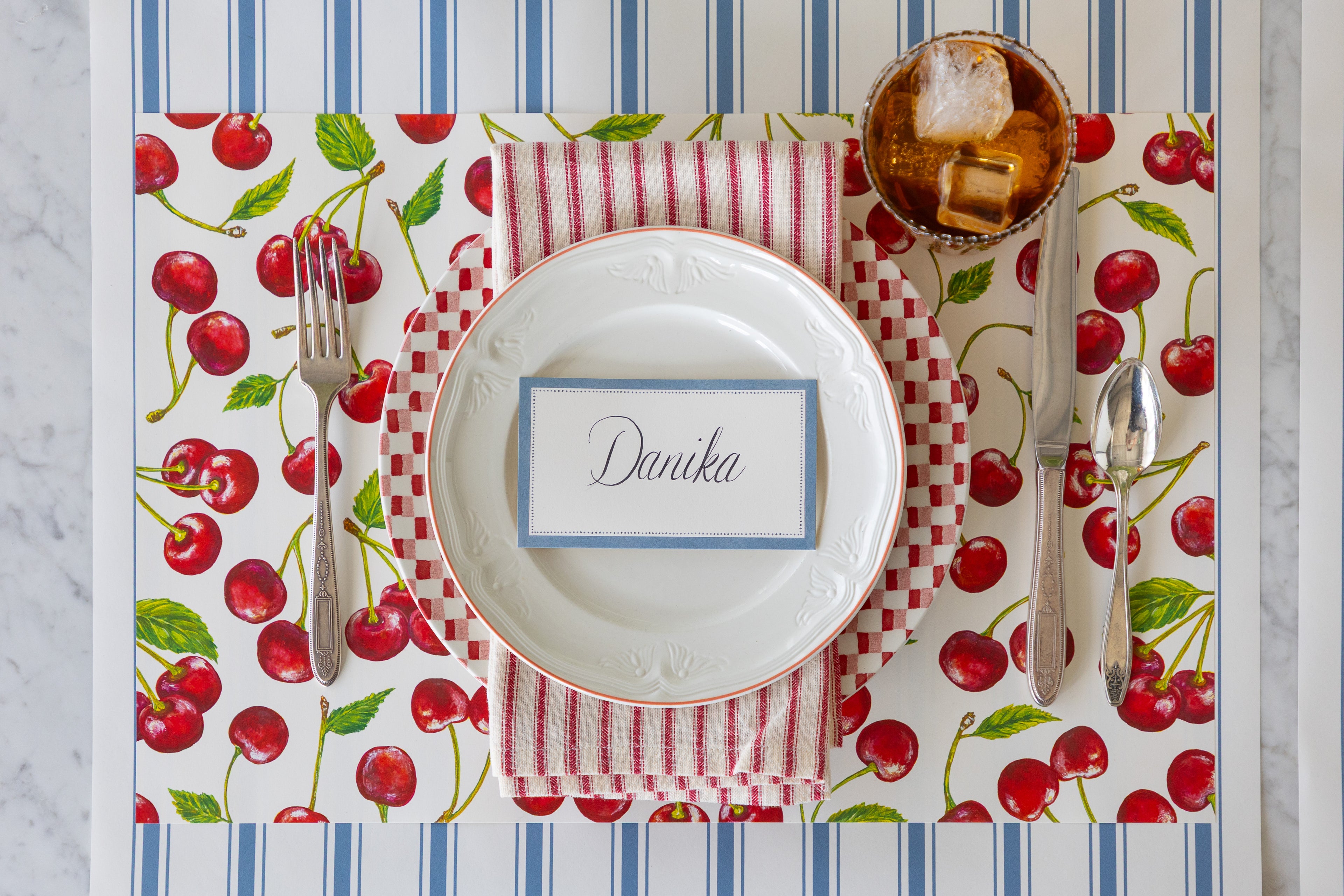 A place setting with the Cherries Placemat, red and white plates and napkin and Cornflower Blue Place Card on the Cornflower Blue Linen Stripe Runner.