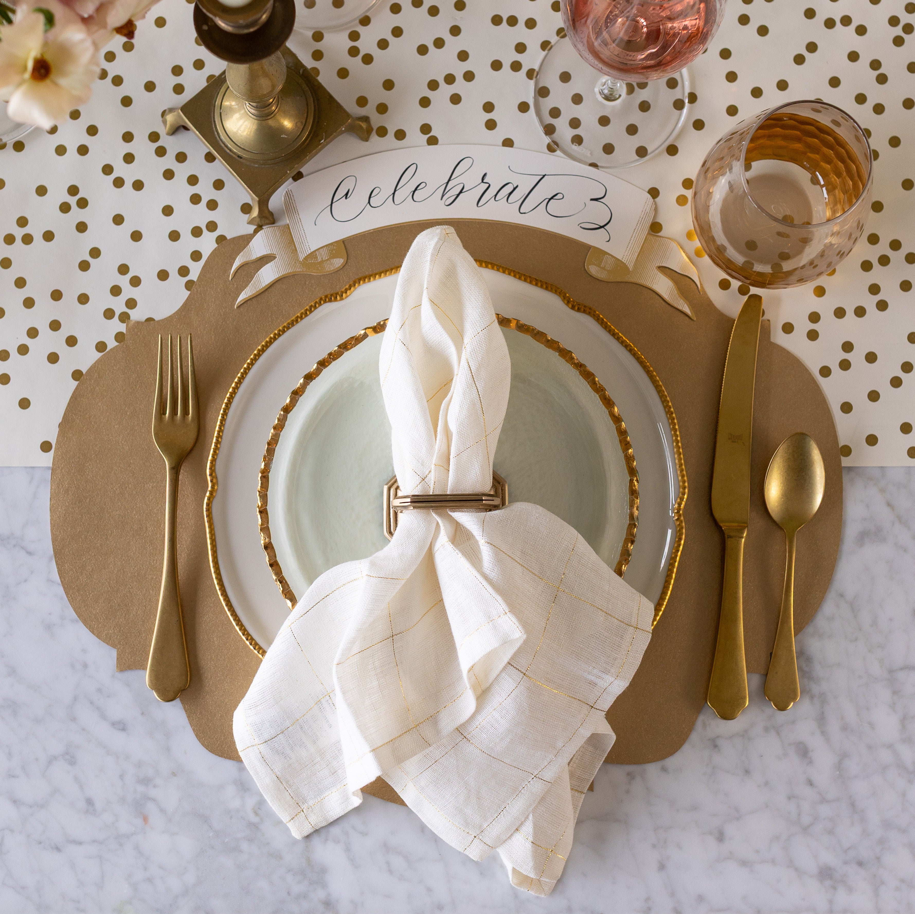 An elegant place setting featuring the Die-cut Gold French Frame Placemat under gold rimmed dinnerware with a napkin in a holder atop, gold flatware, glassware, Gold Banner Table Accent reading "Celebrate" and Gold Confetti Runner underneath.