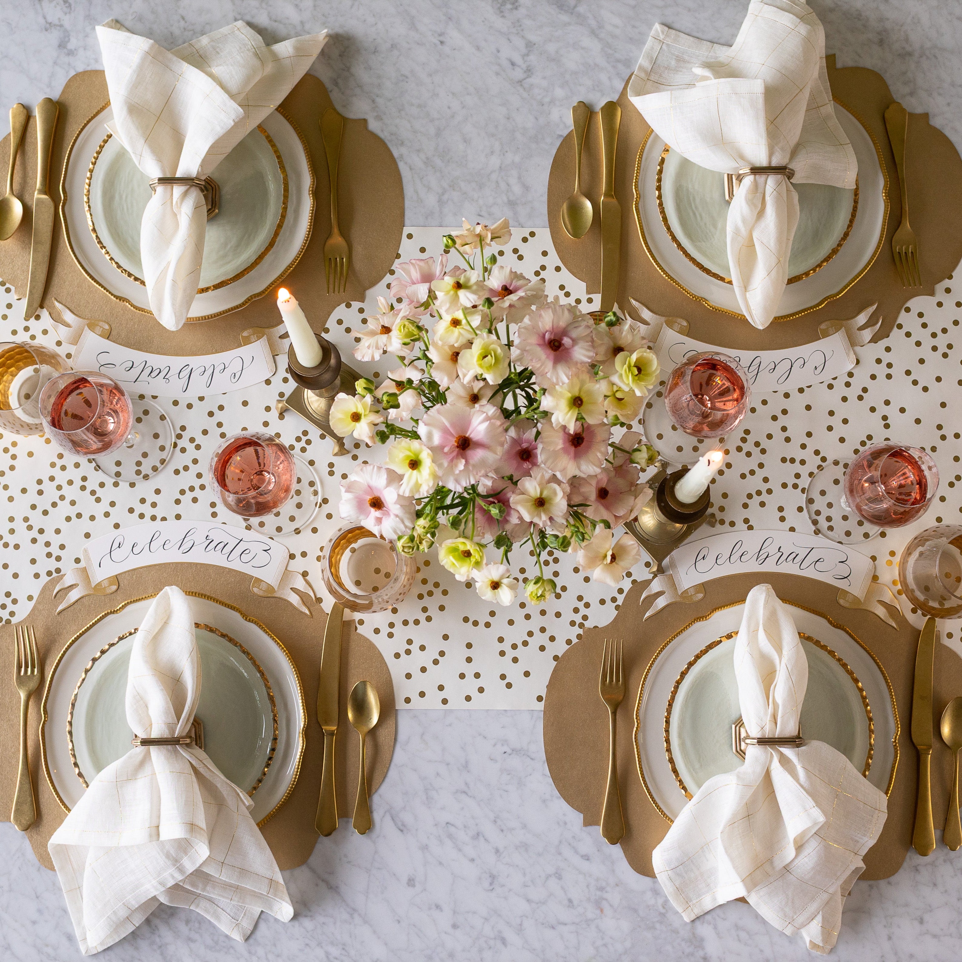 An elegant table setting for four with a floral centerpiece and lit candles, featuring the Die-cut Gold French Frame Placemats with gold rimmed dinnerware, gold flatware, glasses of wine, Gold Banner Table Accents reading "Celebrate" and Gold Confetti Runner underneath.