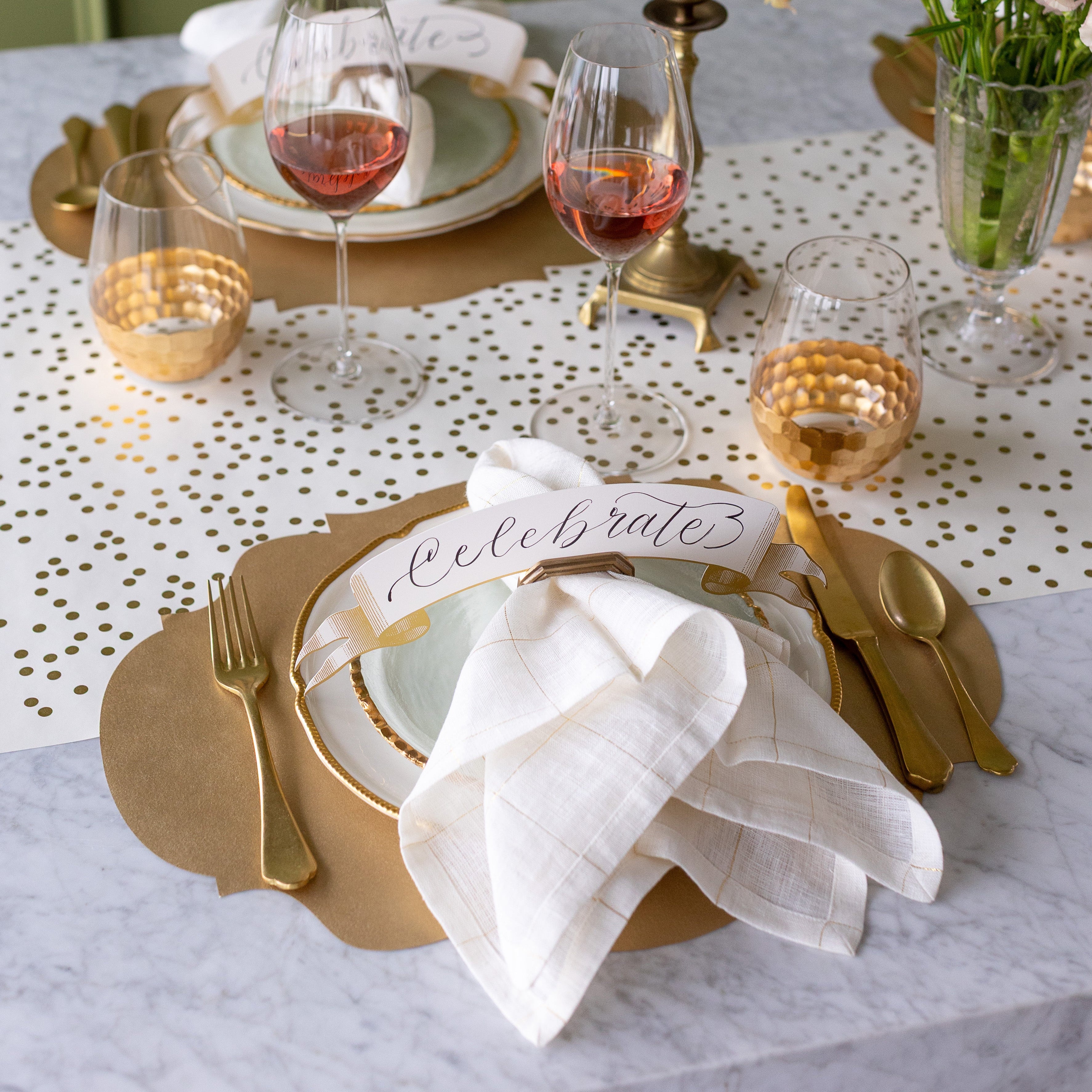 An elegant table setting featuring the Die-cut Gold French Frame Placemat, the Gold Banner Table Accent reading "Celebrate" in the Napkin Ring with Place Card Holder along with a napkin, and Gold Confetti Runner underneath.