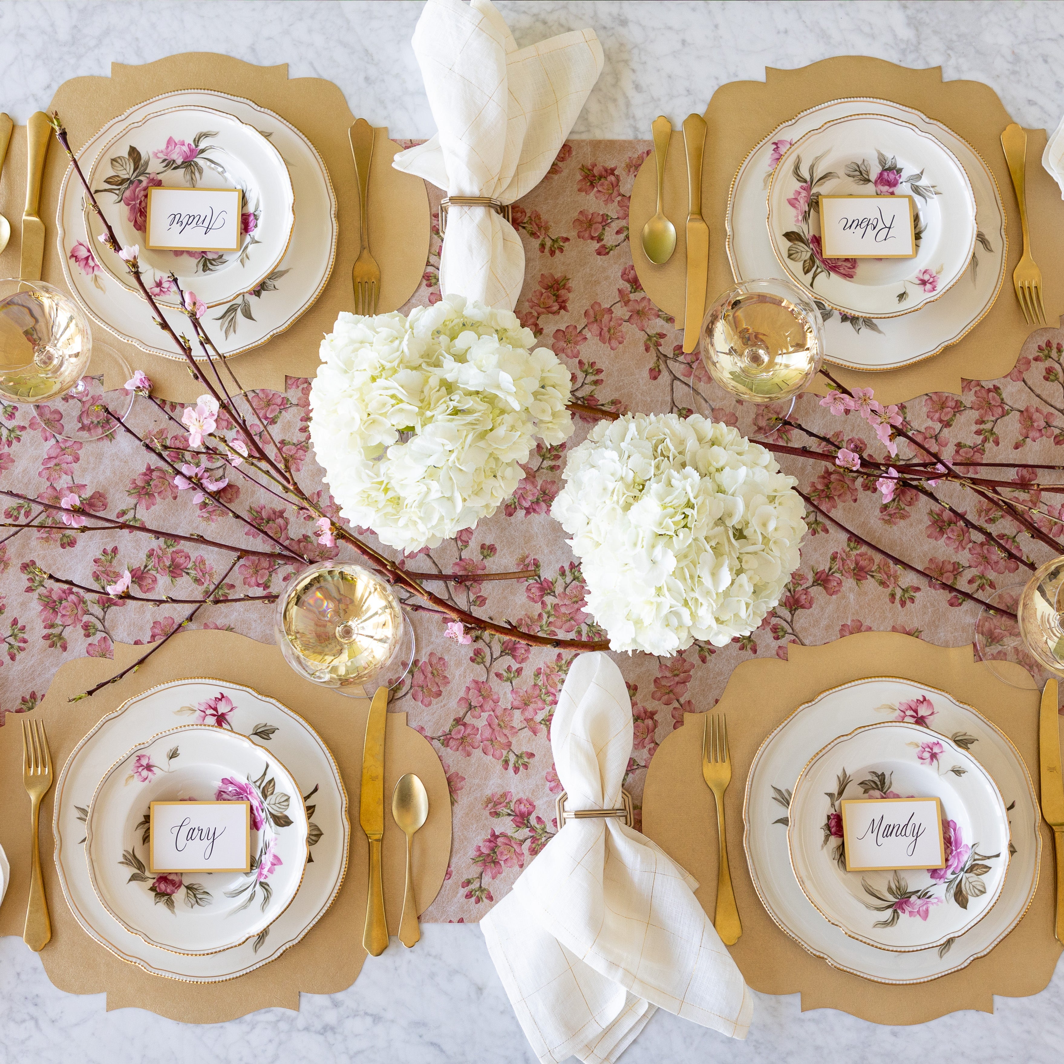 An elegant table setting for four with flowers and cherry blossom branches, featuring the Die-cut Gold French Frame Placemats under vintage floral dinnerware with the Gold Frame Place Cards atop, gold flatware, glasses of wine and the Cherry Blossom Runner underneath.