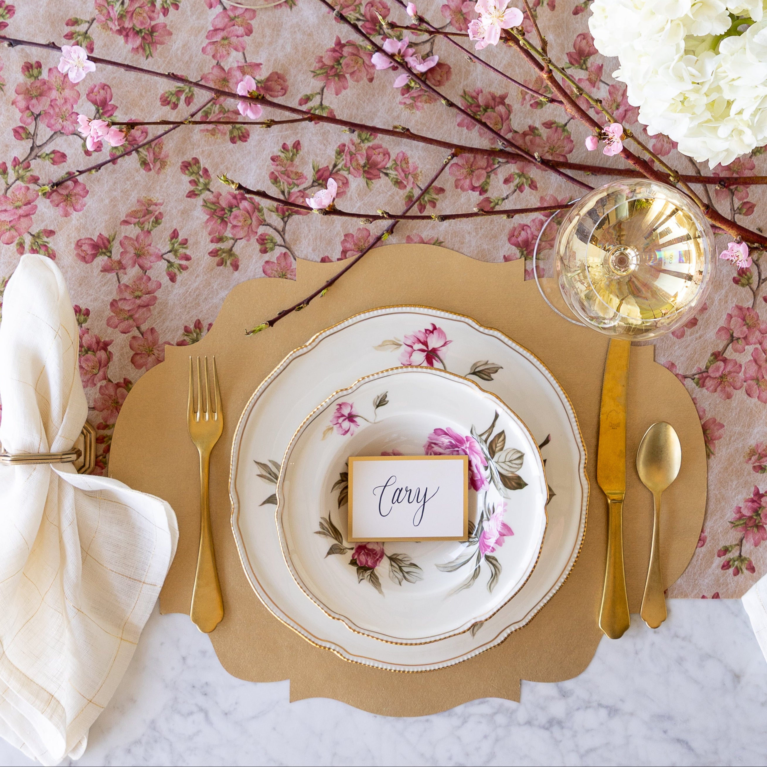 An elegant table setting with flowers and a cherry blossom branch, featuring the Die-cut Gold French Frame Placemat under vintage floral dinnerware with the Gold Frame Place Card reading "Cary" atop, gold flatware, a glass of wine and Cherry Blossom Runner underneath.