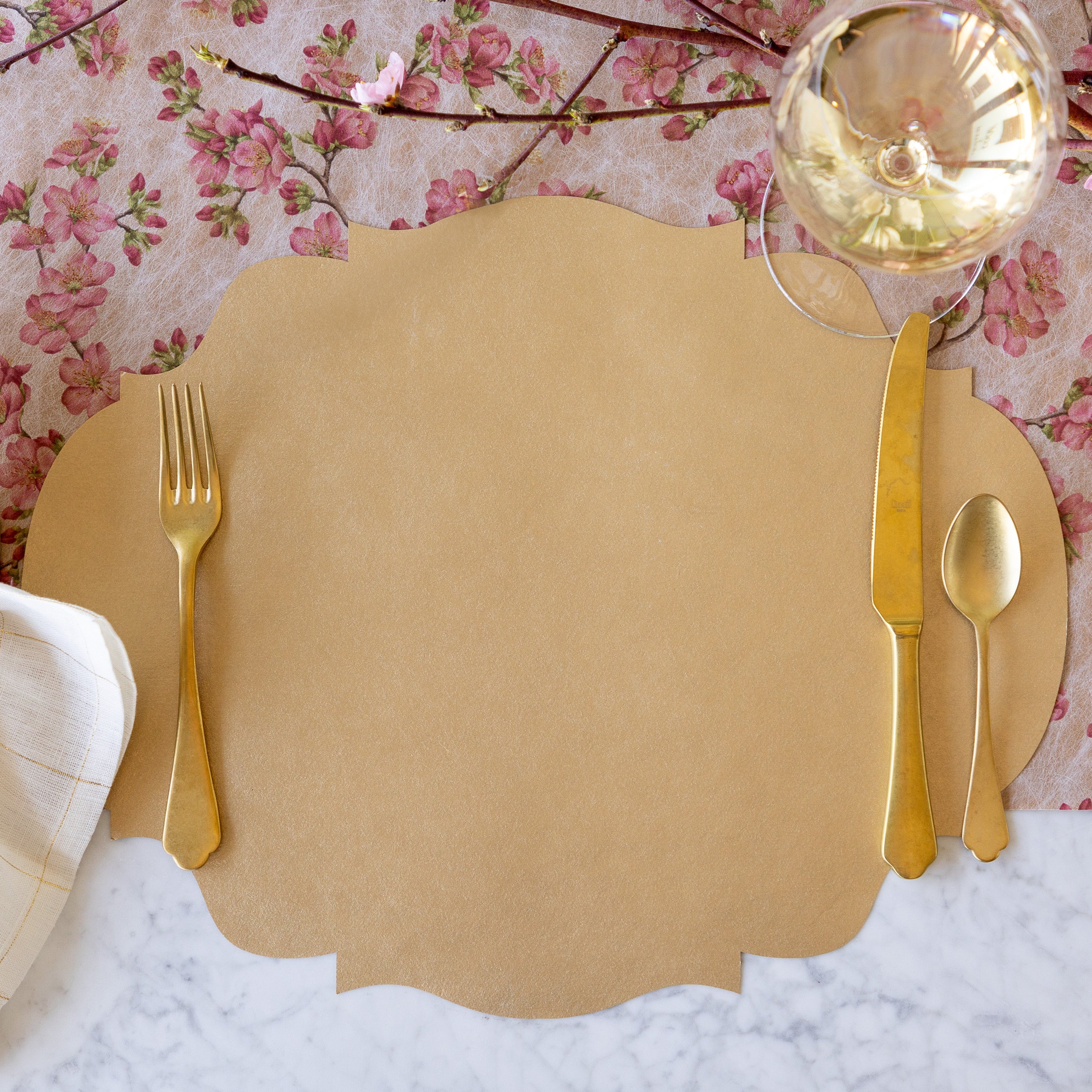 A place setting featuring the Die-cut Gold French Frame Placemat under gold flatware and a glass of wine, with the Cherry Blossom Runner underneath.