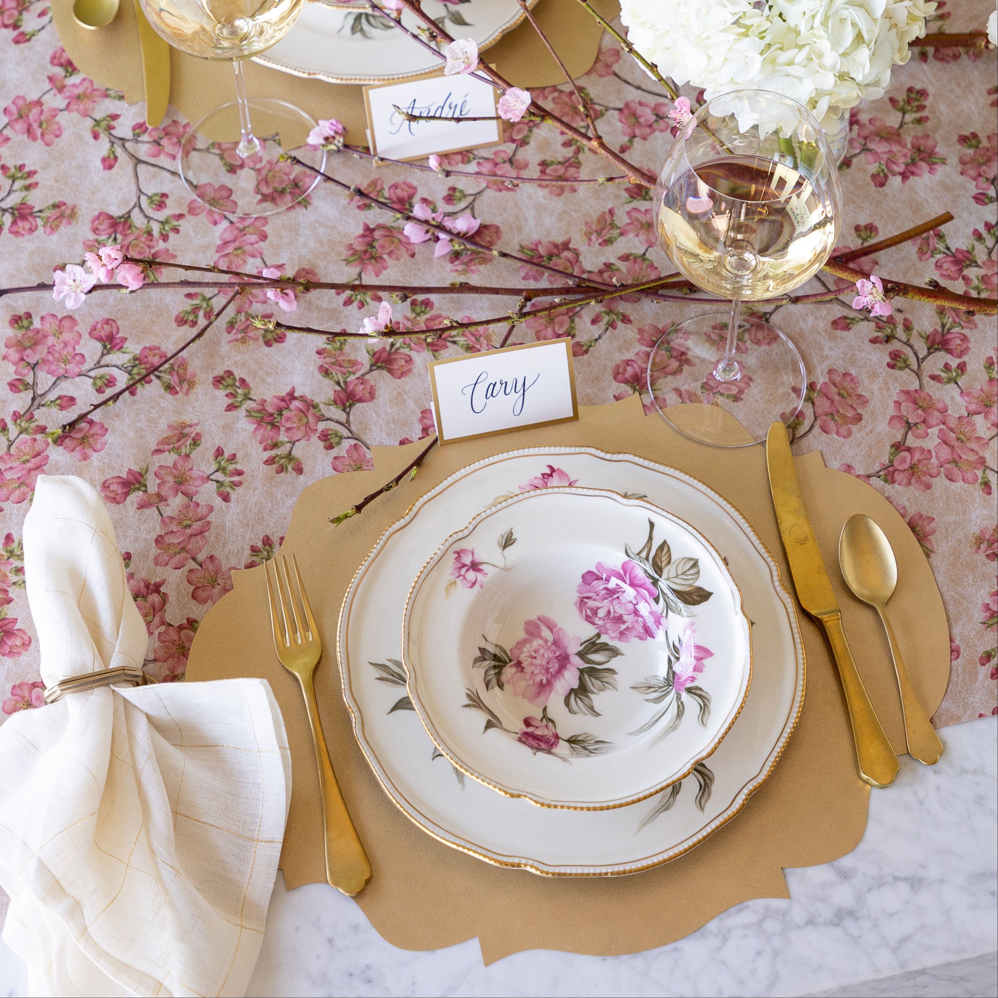 An elegant table setting with flowers and a cherry blossom branch, featuring the Die-cut Gold French Frame Placemat under vintage floral dinnerware, the Gold Frame Place Card reading "Cary", gold flatware, a glass of wine and Cherry Blossom Runner underneath.