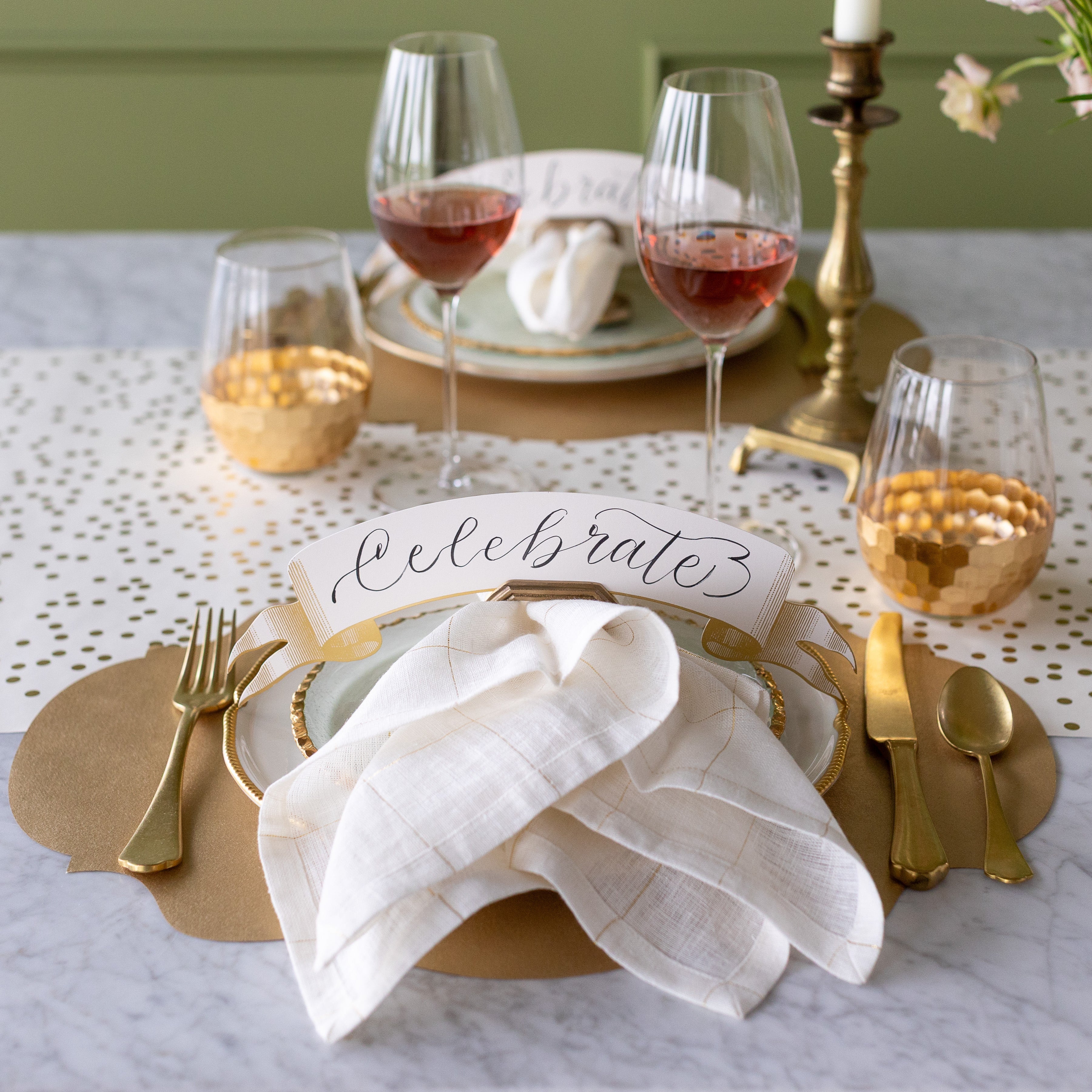 An elegant table setting featuring the Die-cut Gold French Frame Placemat with the Classic Gold Banner Table Accent reading "Celebrate" held by the Napkin Ring with Place Card Holder, atop the dinner plates.