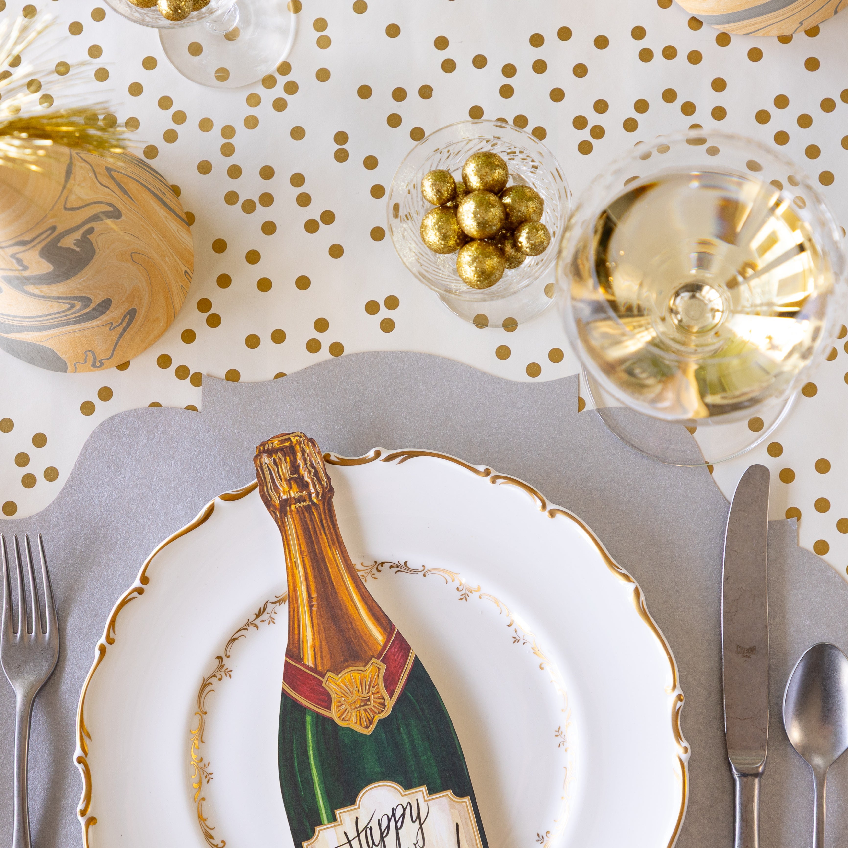 An elegant place setting featuring the Die-cut Silver French Frame Placemat under a gold rimmed dinner plate with the Champagne Table Accent atop, a glass of wine, cup of glittery gold balls, a marbled party hat, and Gold Confetti Runner underneath.