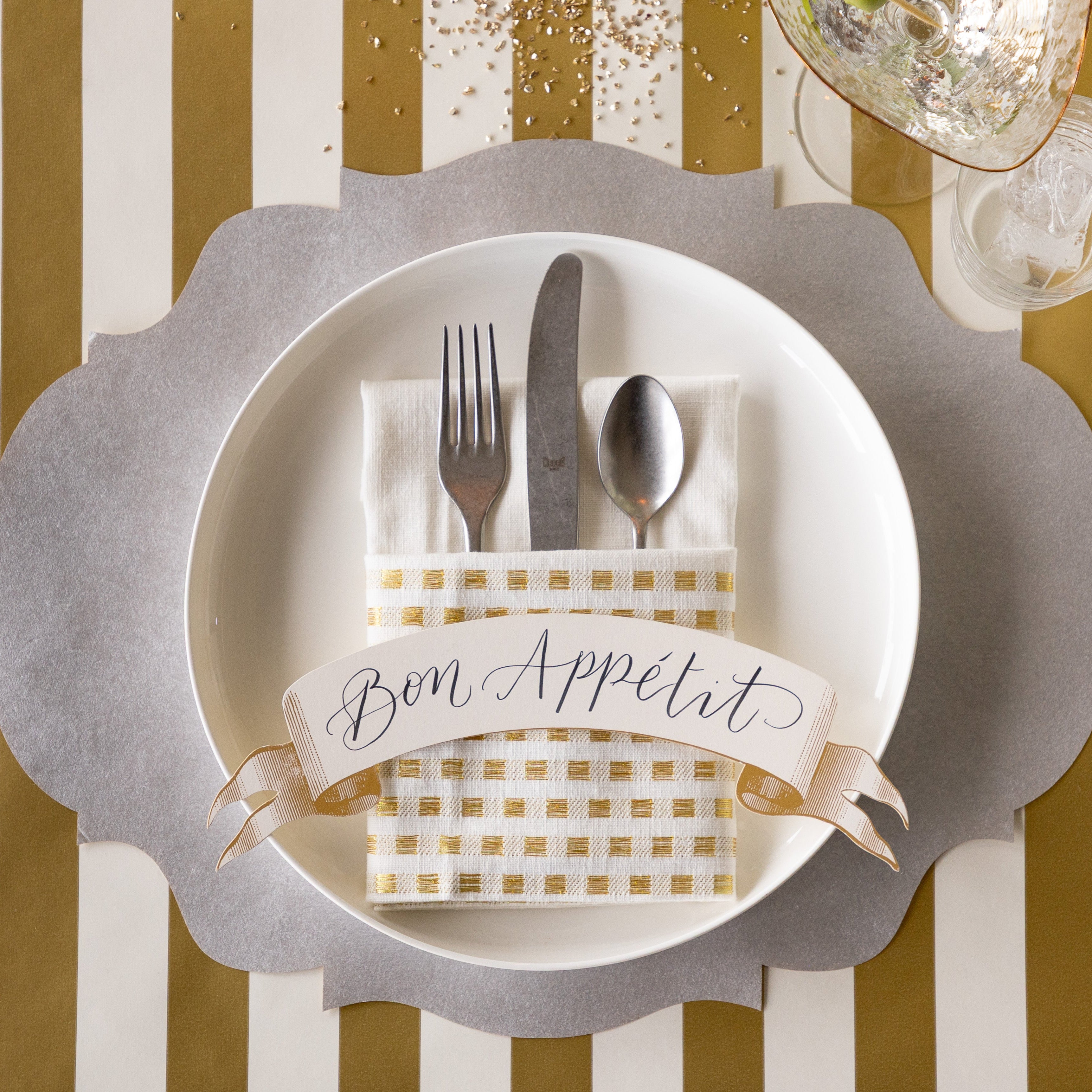 A place setting featuring the Die-cut Silver French Frame Placemat under a white dinner plate with silverware inside of a folded napkin and Classic Gold Banner Table Accent reading "Bon Appetit" atop and the Gold Classic Stripe Runner underneath.
