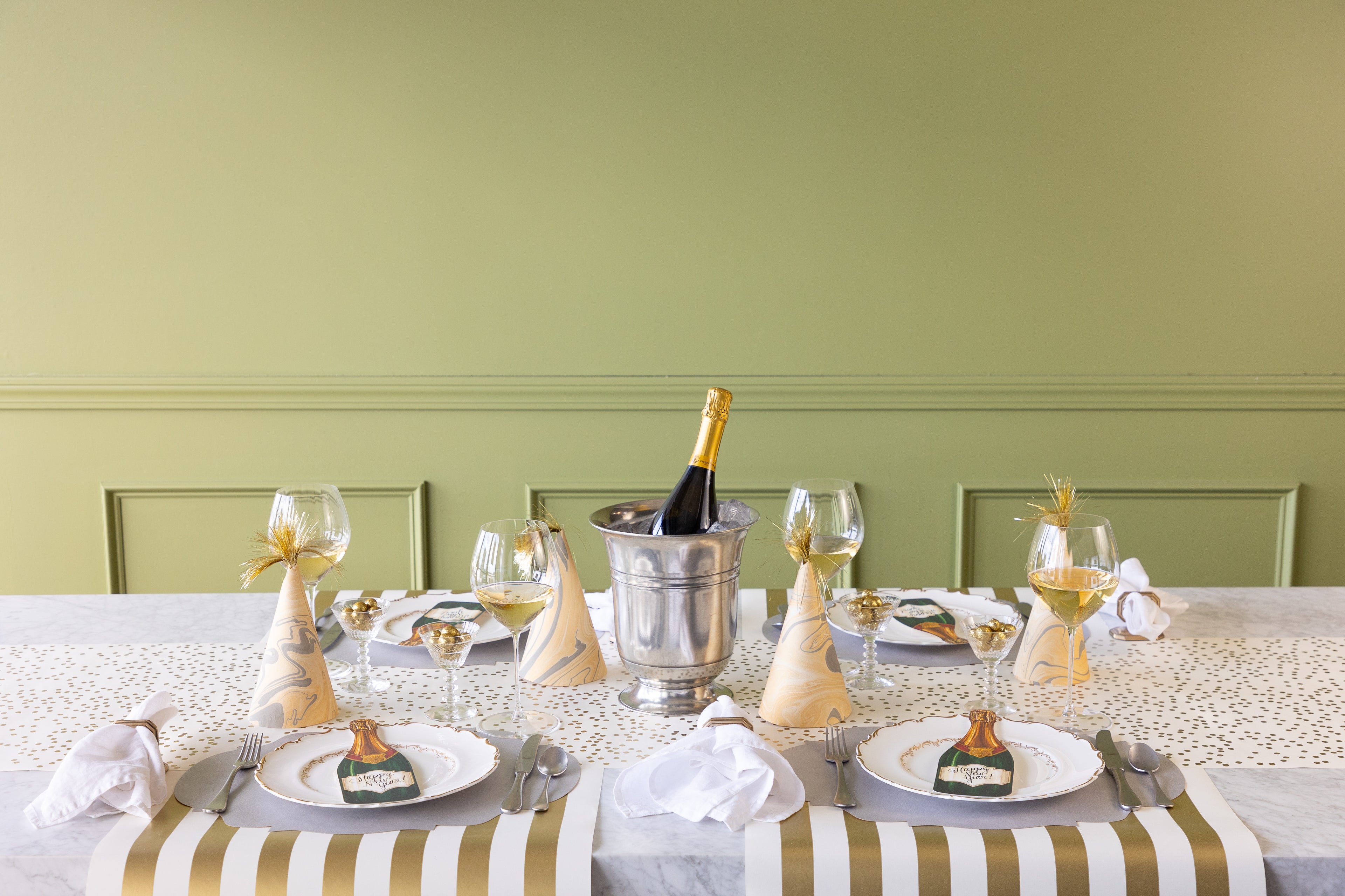 A festive table with the confetti runner on it and party hats at 4 place settings. White plates and a bottle of champagne are also on the table.