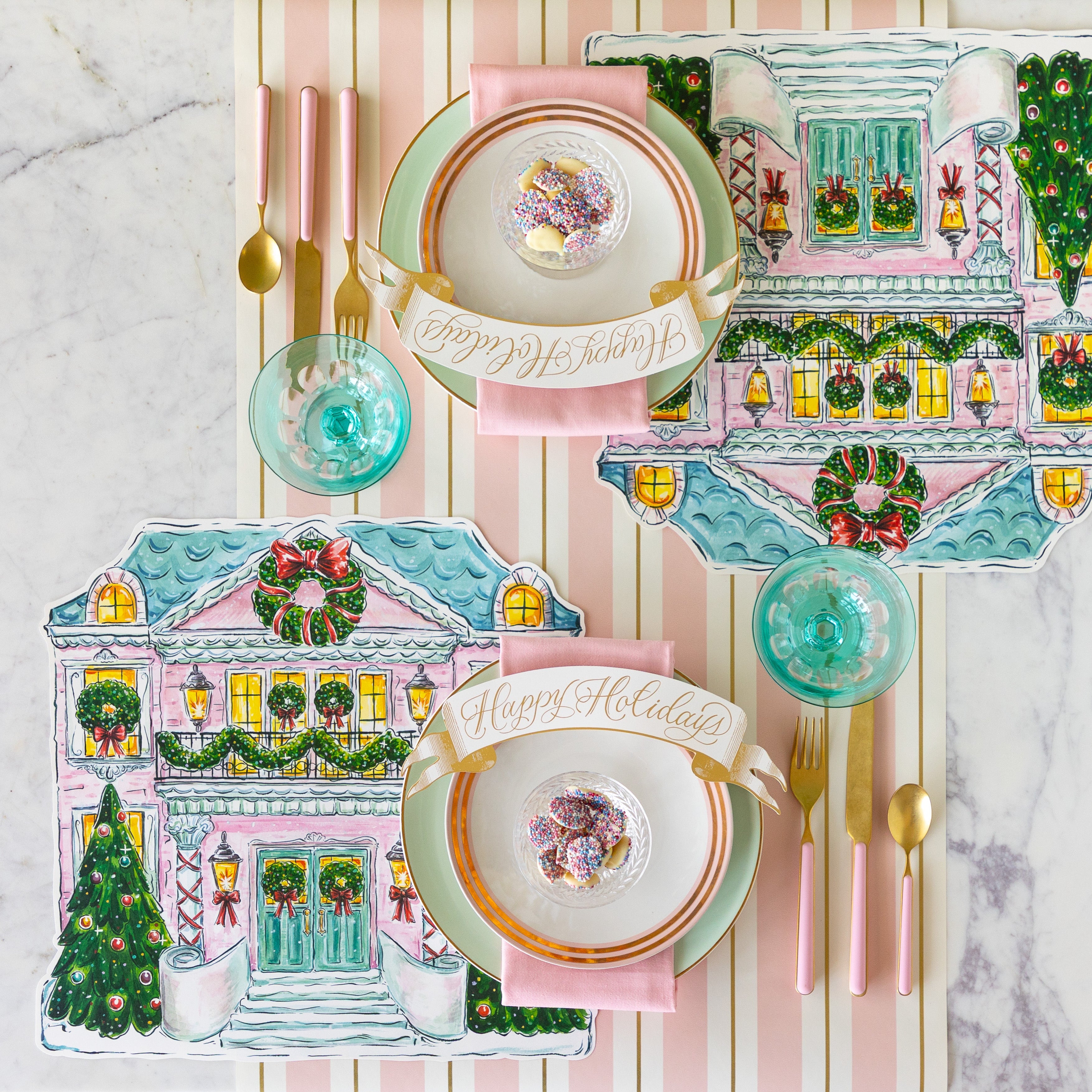 A table set with the Die-cut Holiday Home Placemats, seafoam and gold-rimmed plates, pink napkins, bowl of white chocolate buttons and Happy Holidays Banner Table Accent atop, and pink flatware on the Pink & Gold Awning Stripe Runner.