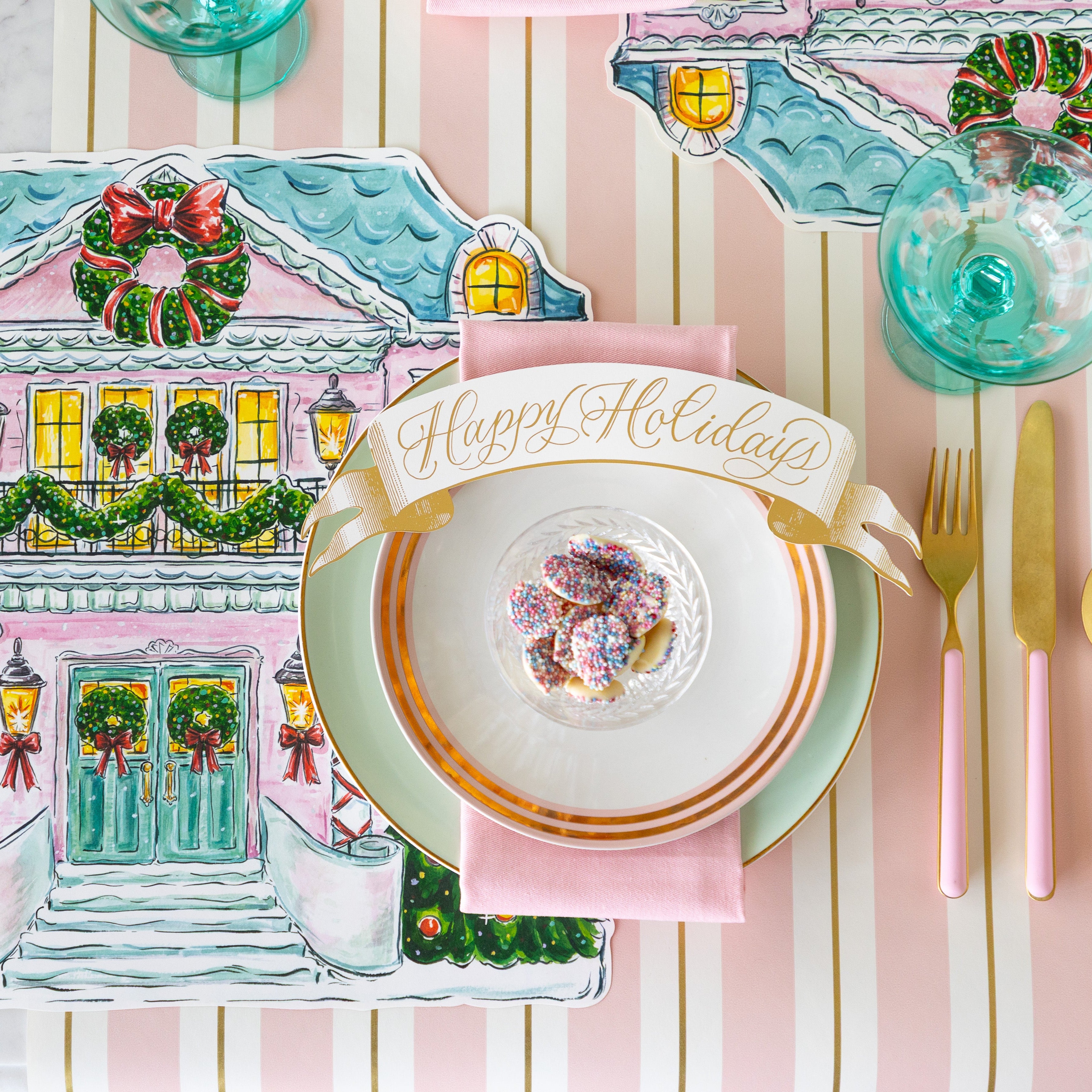 Happy Holidays Banner Table Accent atop a place setting with gold rimmed plates, bowl of chocolate buttons, a pink napkin, Die-cut Home for the Holidays Placemat and Pink & Gold Awning Stripe Runner underneath.